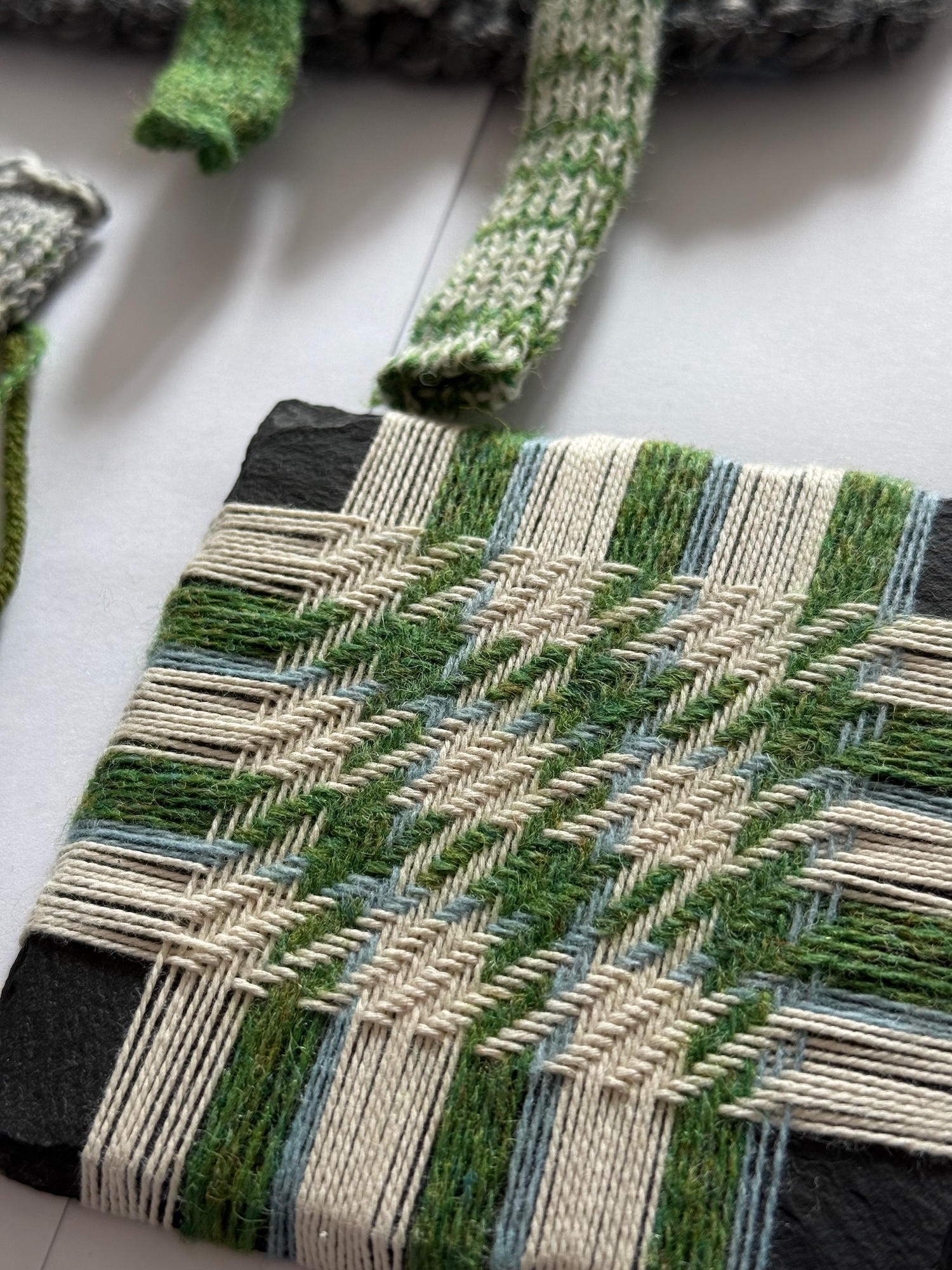 Weaving in progress on loom showing green, beige and blue striped pattern with diagonal chevron texture in woven fabric.