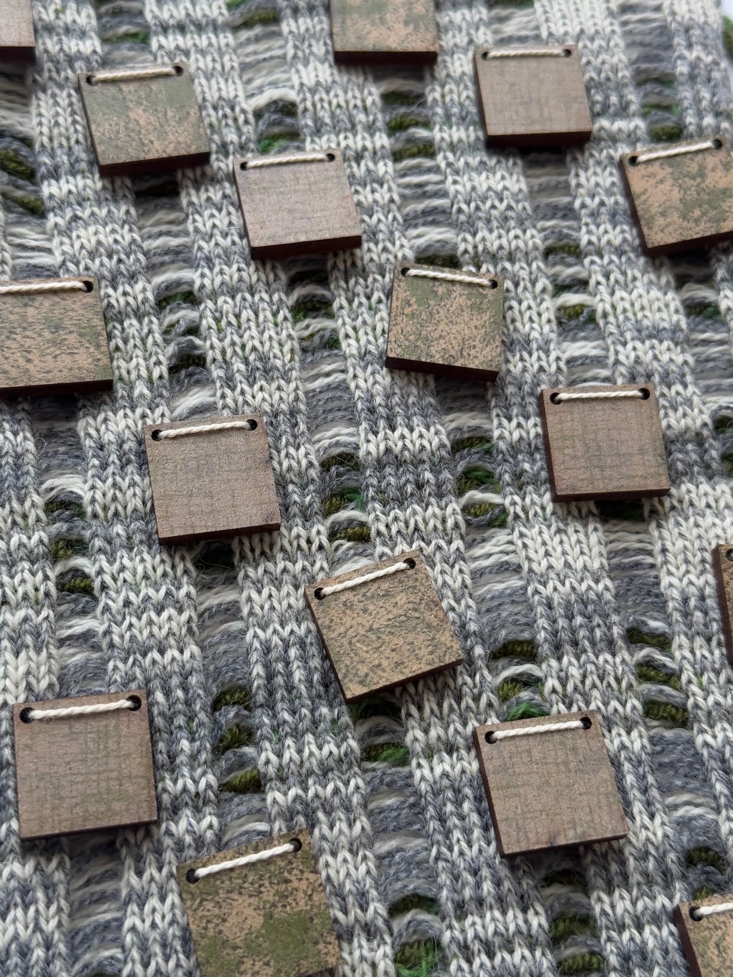Grey knitted fabric with vertical cable pattern interwoven with brown rectangular leather or cardboard patches attached with metal rings.