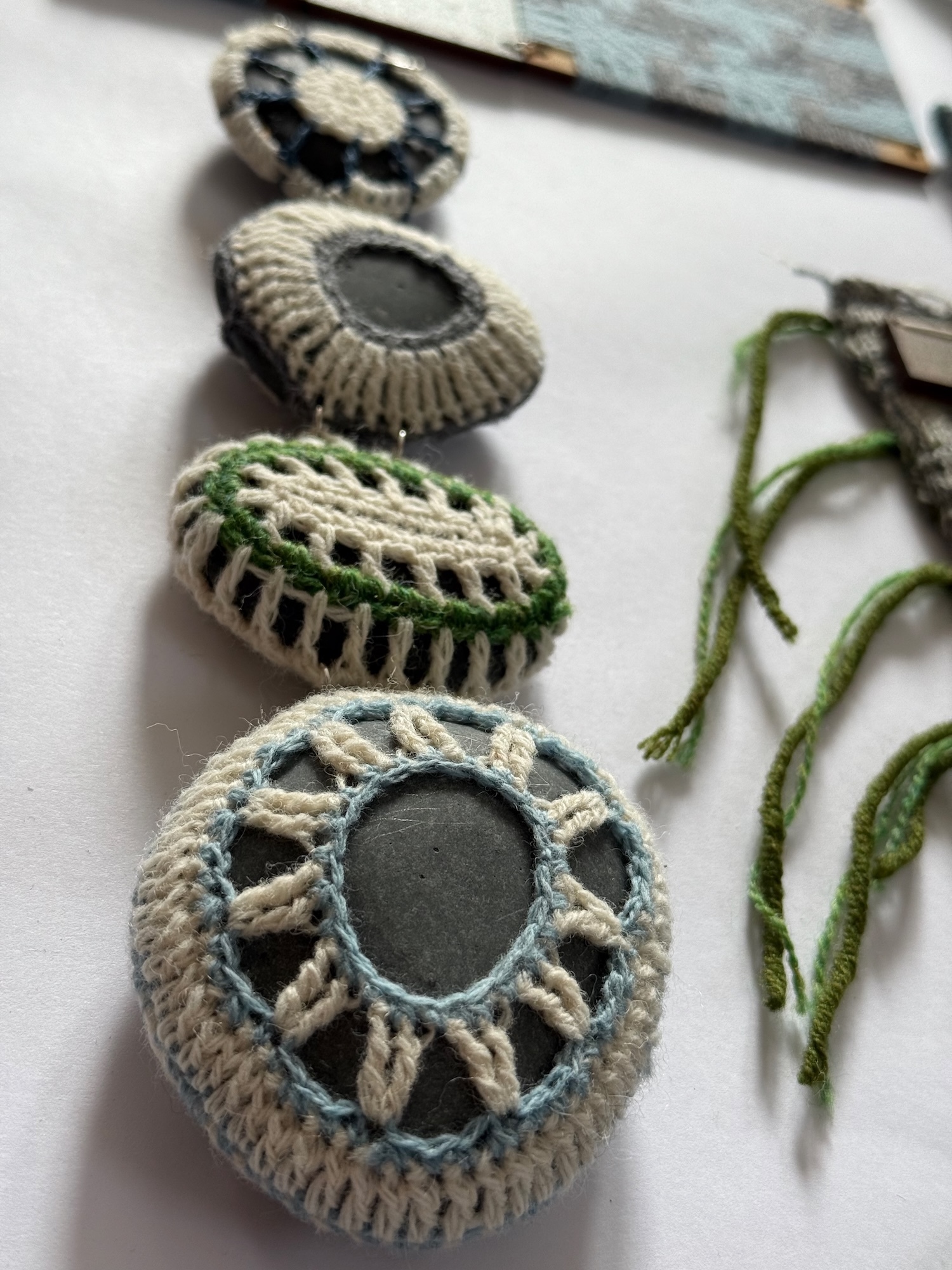 Four crocheted stone covers in grey, green and blue yarn with decorative patterns, arranged vertically on white surface with yarn strands.