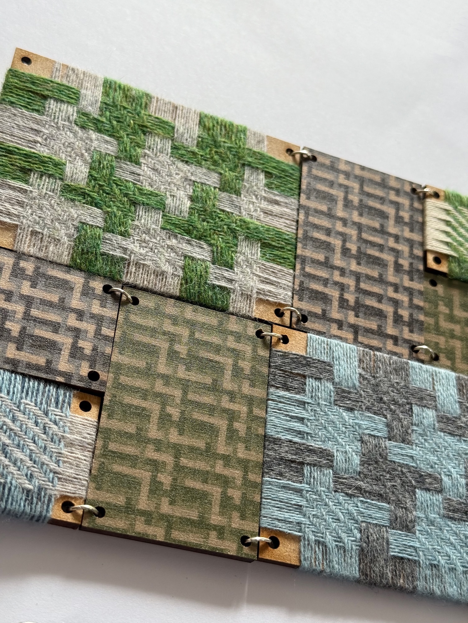 Woven textile samples in green, blue, brown, and cream colours arranged in rectangular grid pattern with metal rings attached.