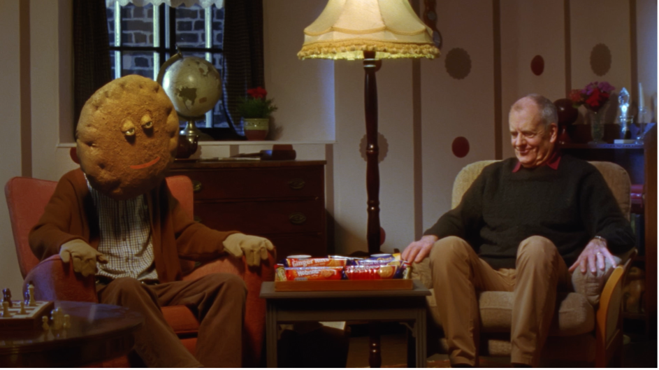 Still from BA (Hons) Film Production student commercial "Baz and Biscuit", showing two people sitting in armchairs with a coffee table and large lamp in between them. The person on the left has a biscuit for a head.