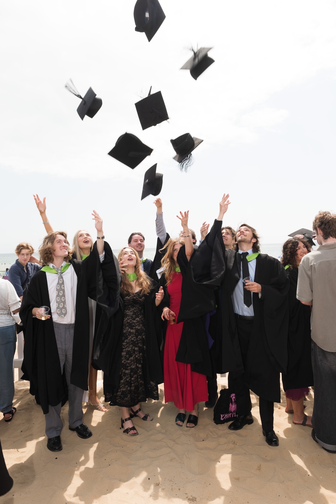 Group of graduates in black gowns and mortarboards throwing their caps into the air, arms raised upwards against white sky.