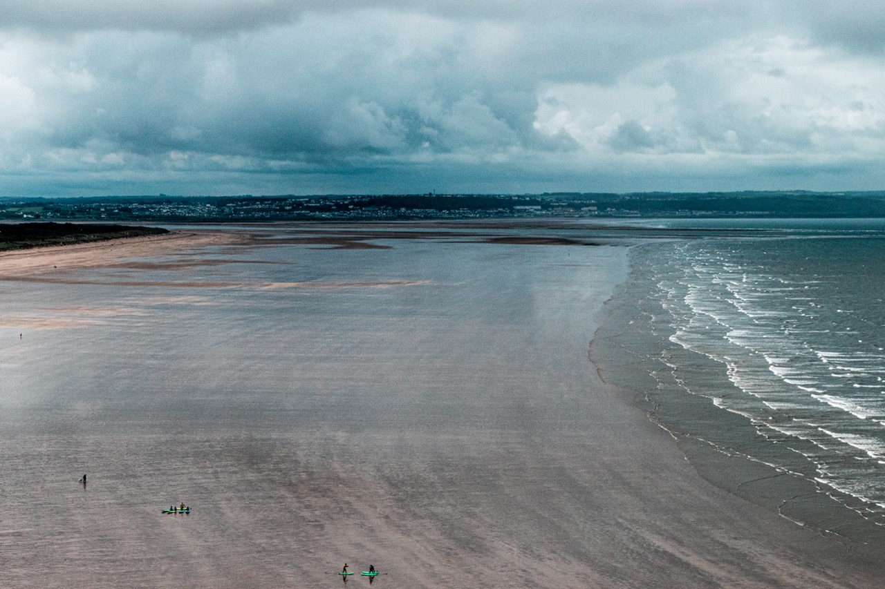 A wide sandy beach with gentle waves on the right and a cloudy sky above, viewed across a distant shoreline.