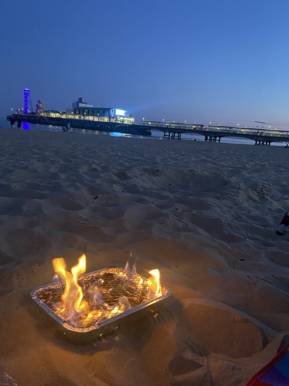Disposable barbecue with flames burning on wet sand at dusk, illuminated pier with purple lights extending into calm sea under blue sky.