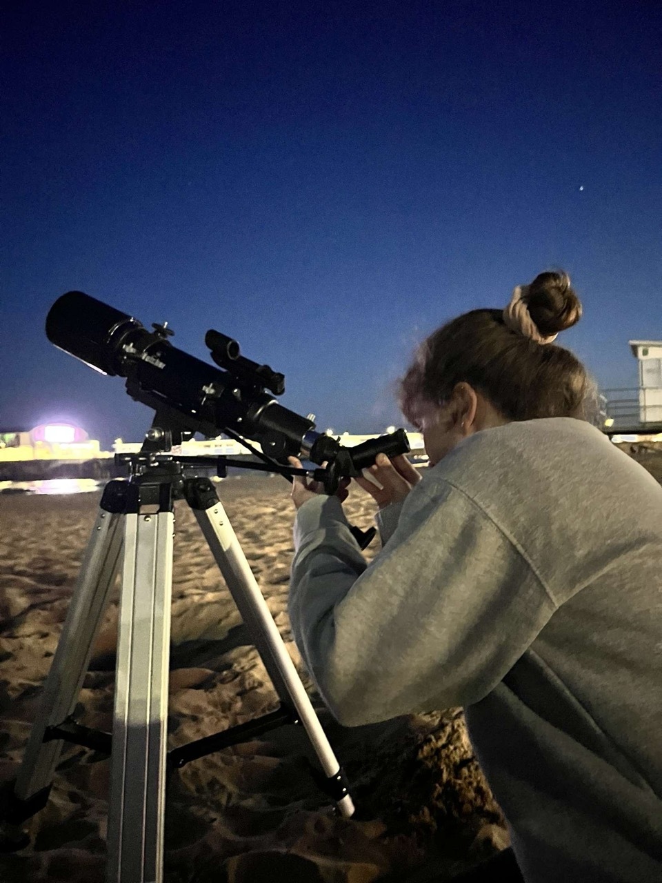 Woman in beige jumper looking through black telescope on tripod at twilight, with deep blue sky and distant lights visible.