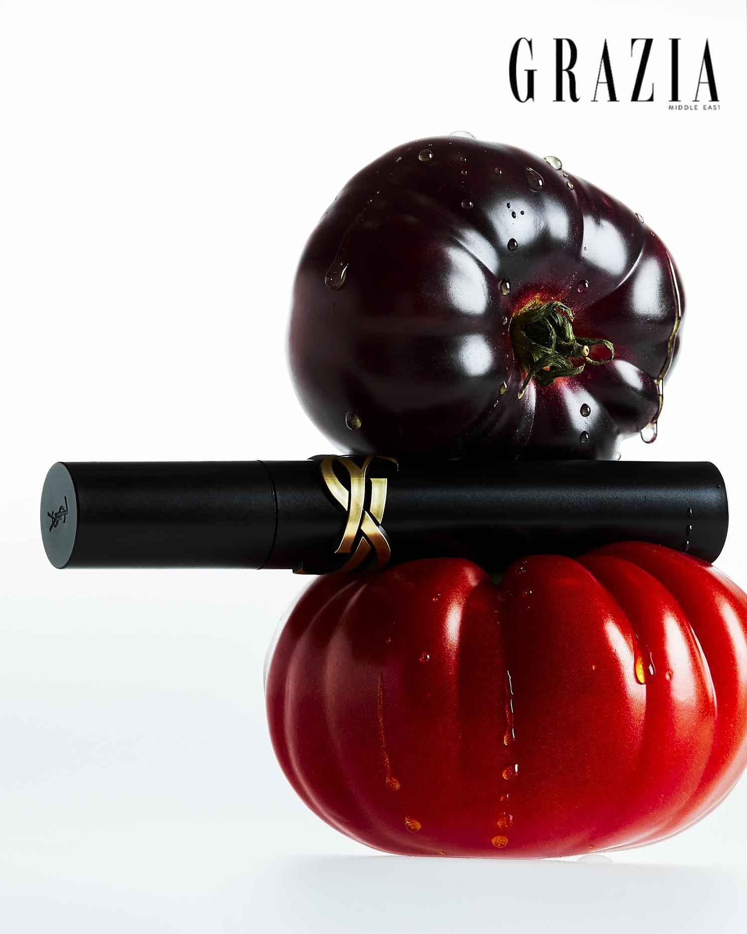 A glossy black lipstick tube positioned between a round red tomato and a dark purple eggplant, both displaying drops of water. The background is white, and the top left corner features the word 'Grazia' in bold letters.