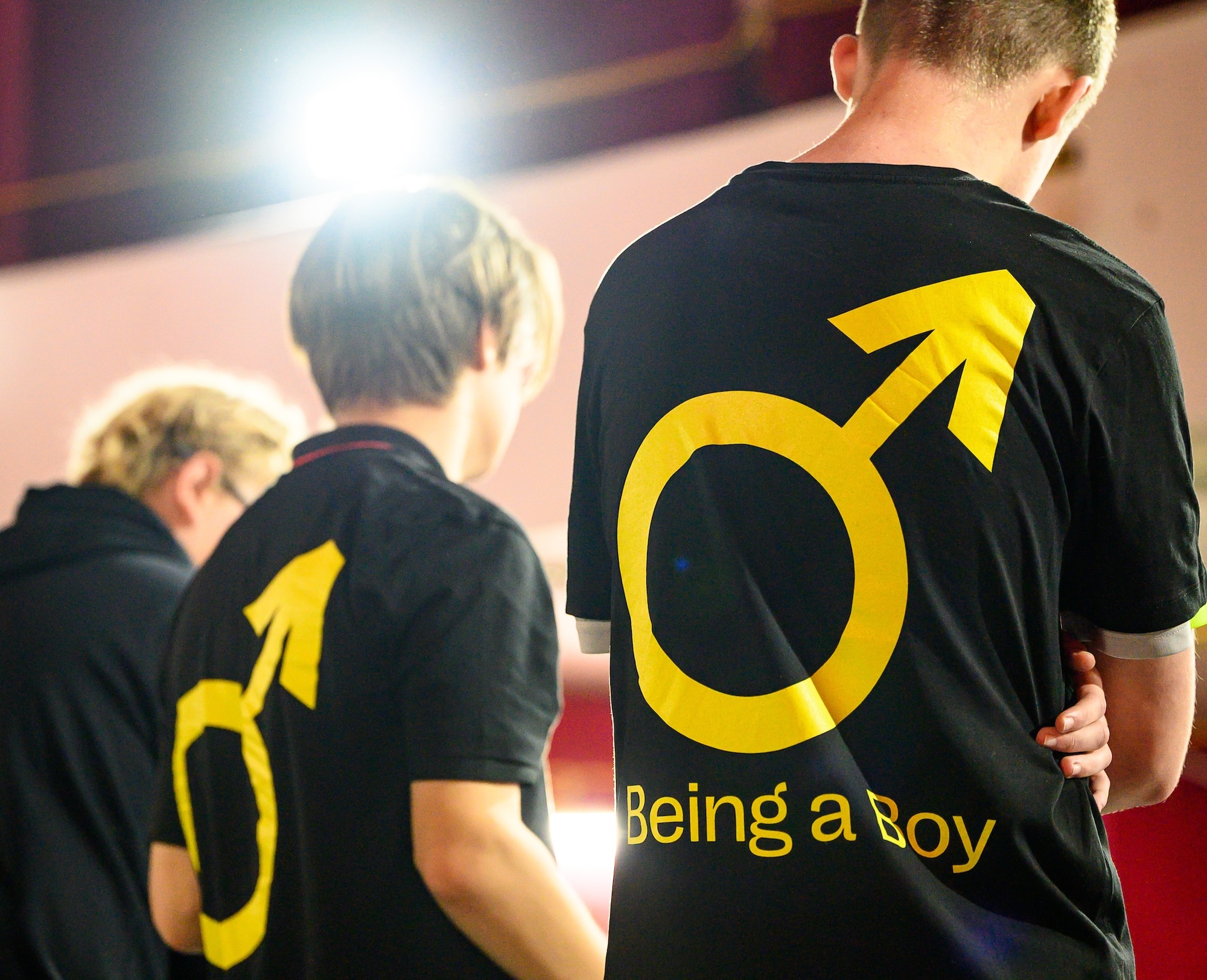 Two young people viewed from the back wearing black T-shirts, which have a yellow male symbol and the words "Being a Boy" underneath printed on them.