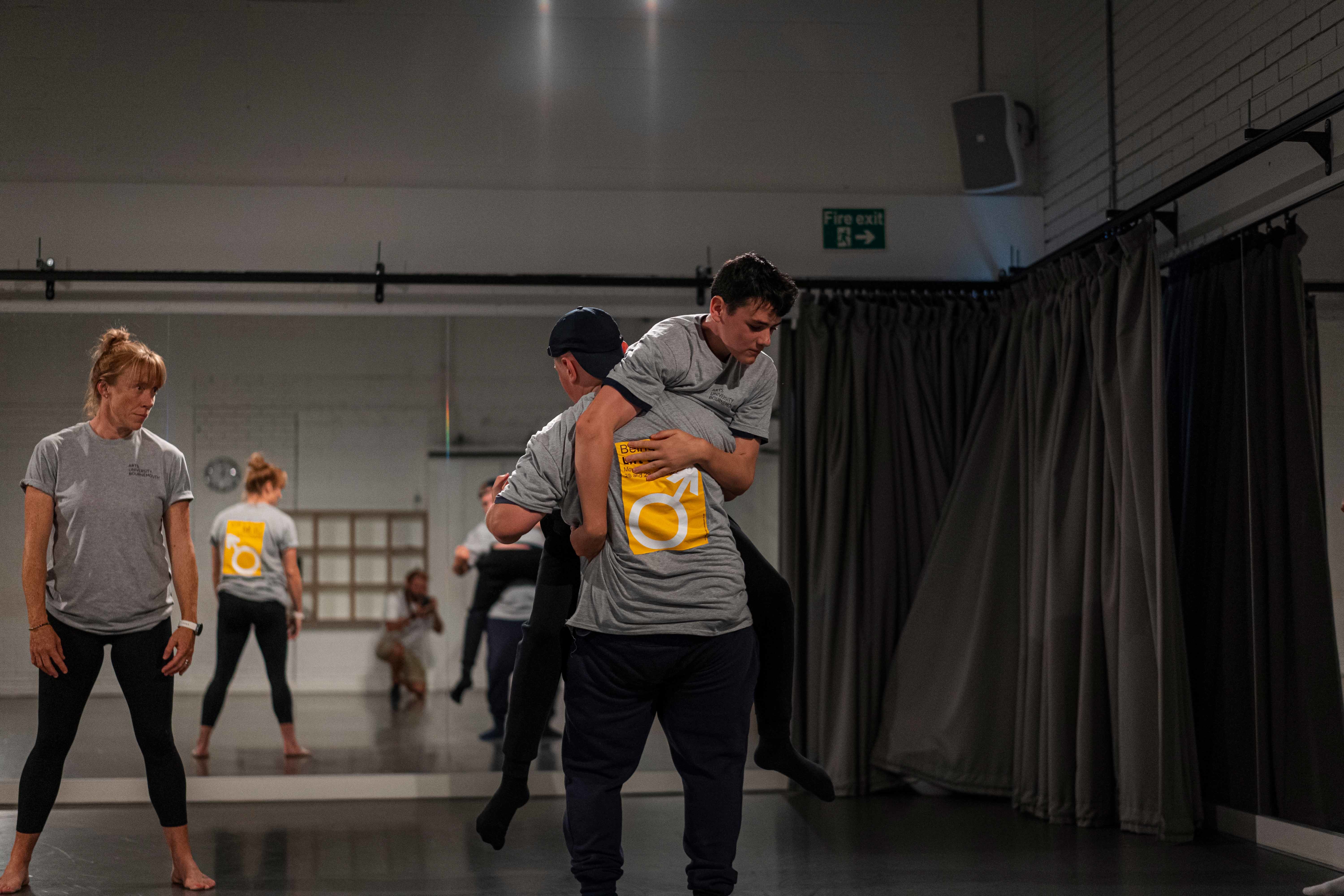 2 students and a dance teacher wearing grey t-shirts with Being a Boy written on the back in a dance studio. The 2 boys are doing lifts.