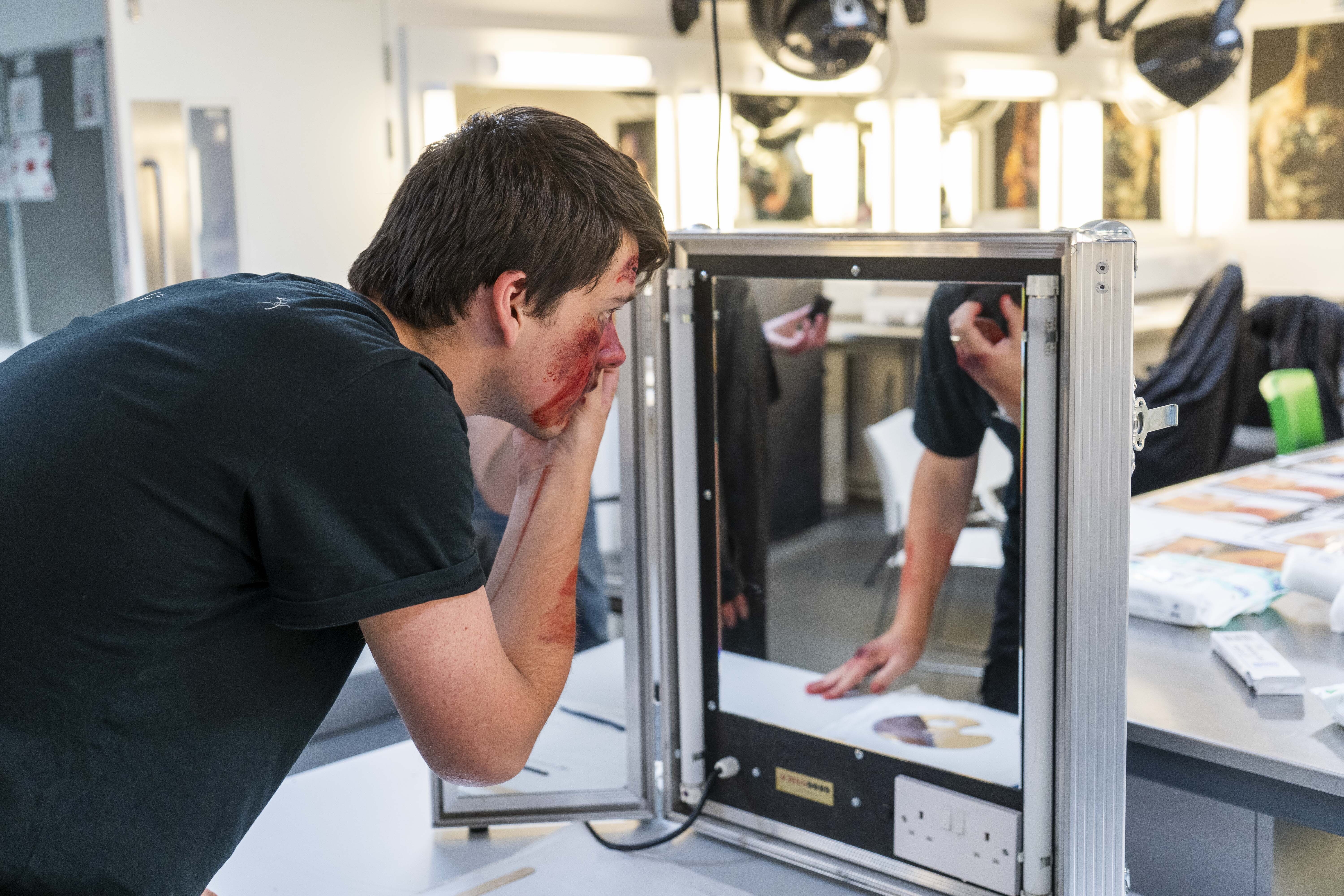 You man applying special effects makeup looking into a light up mirror