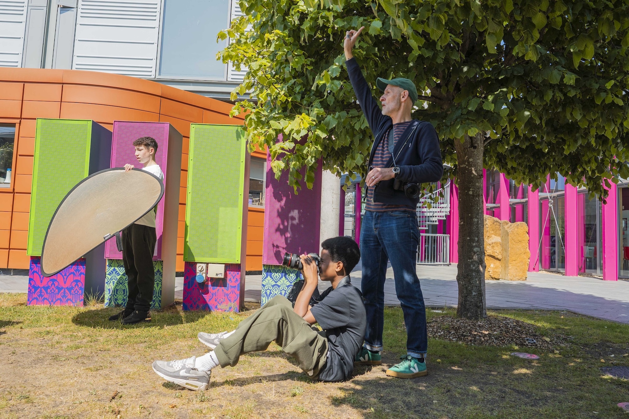 A tutor guiding two young men, one holding a camera, and one holding a light reflector by colourful street mural with bright green, pink, orange and purple panels.