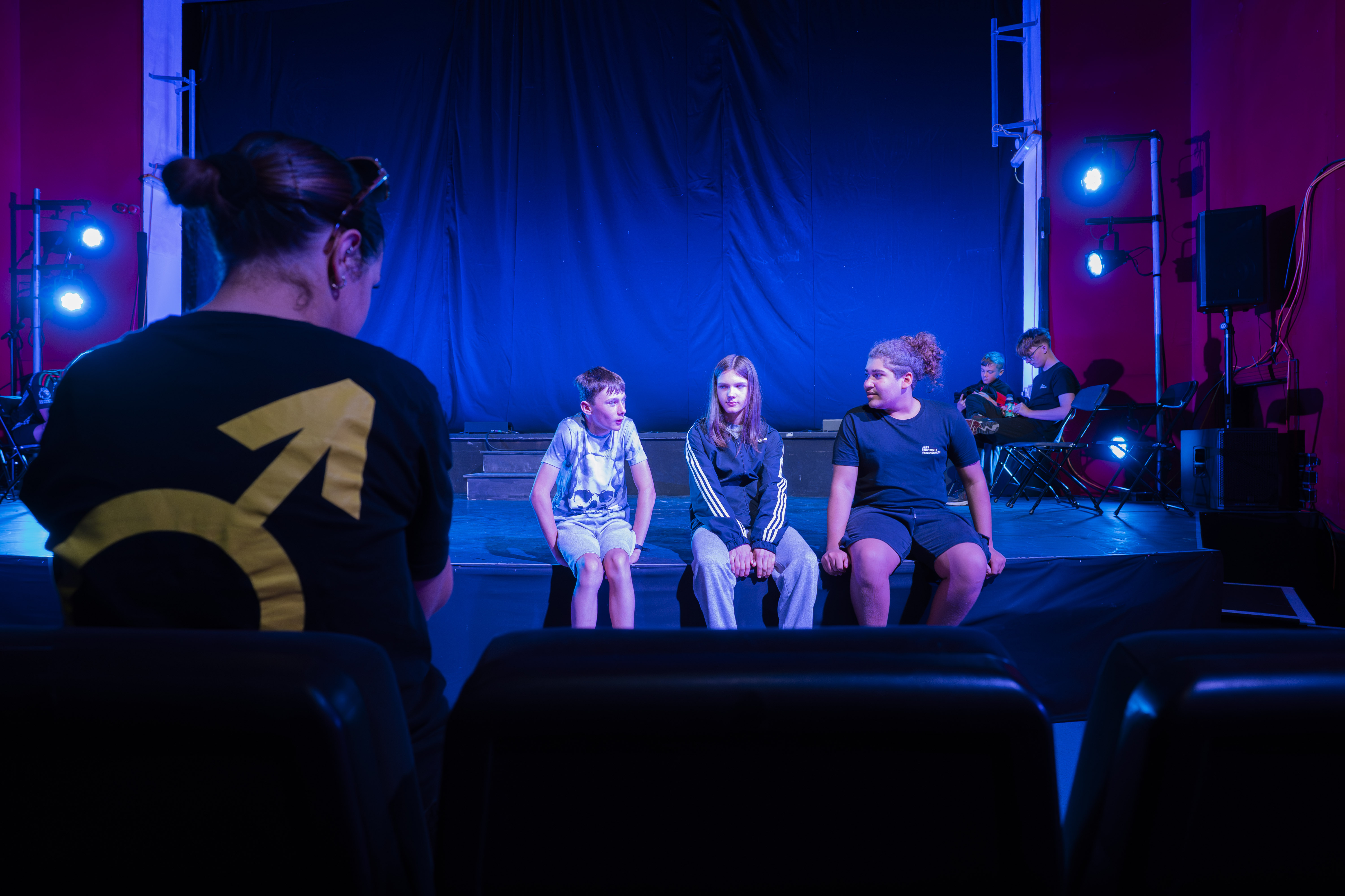 Three young men sitting on stage with chairs under blue and purple lighting in the background and with a member of the Being a Boy team watching the performance practise