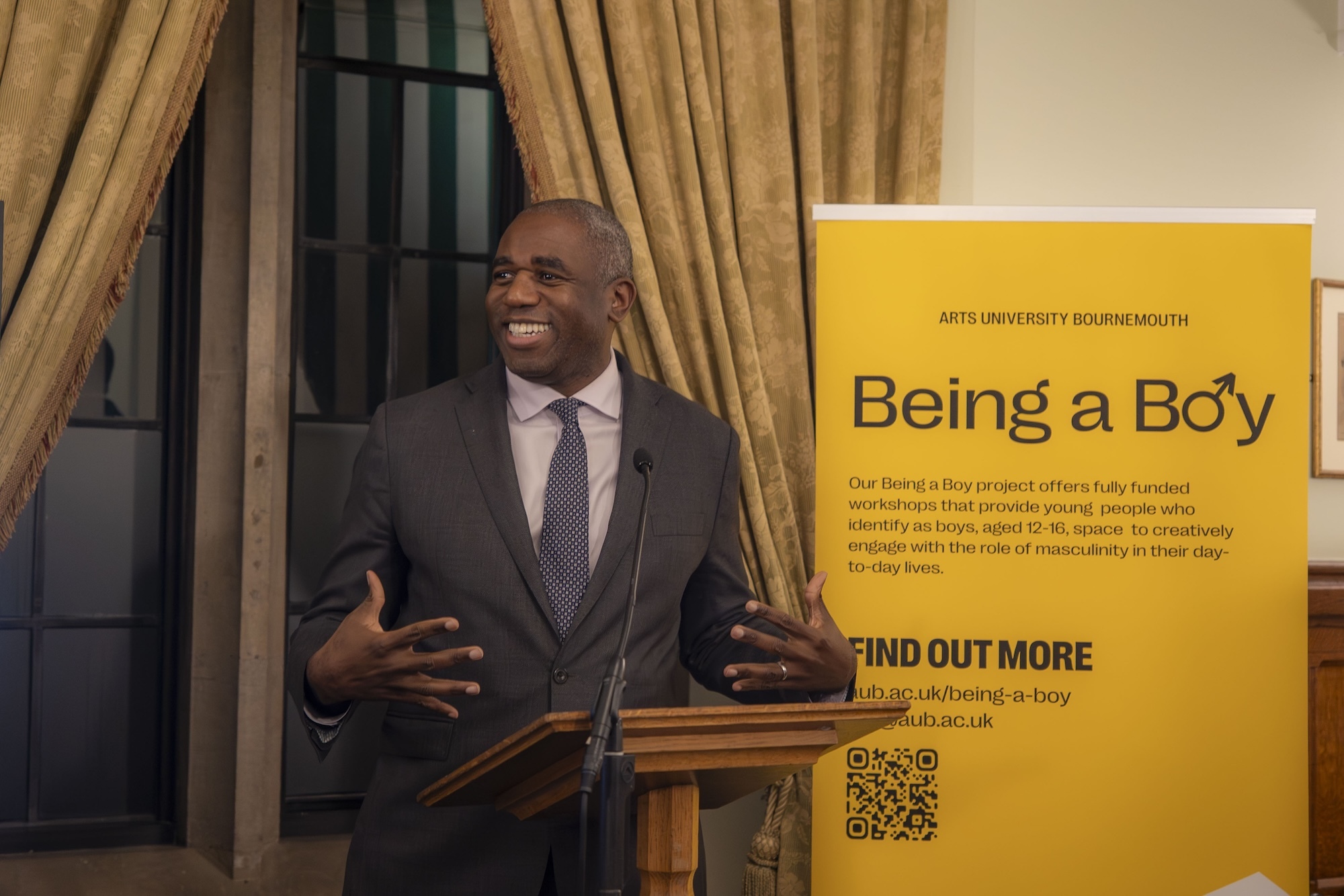 Black man in dark suit speaking at wooden podium, hands gesturing. Yellow banner reads "Being a Boy" beside him. Gold curtains behind.