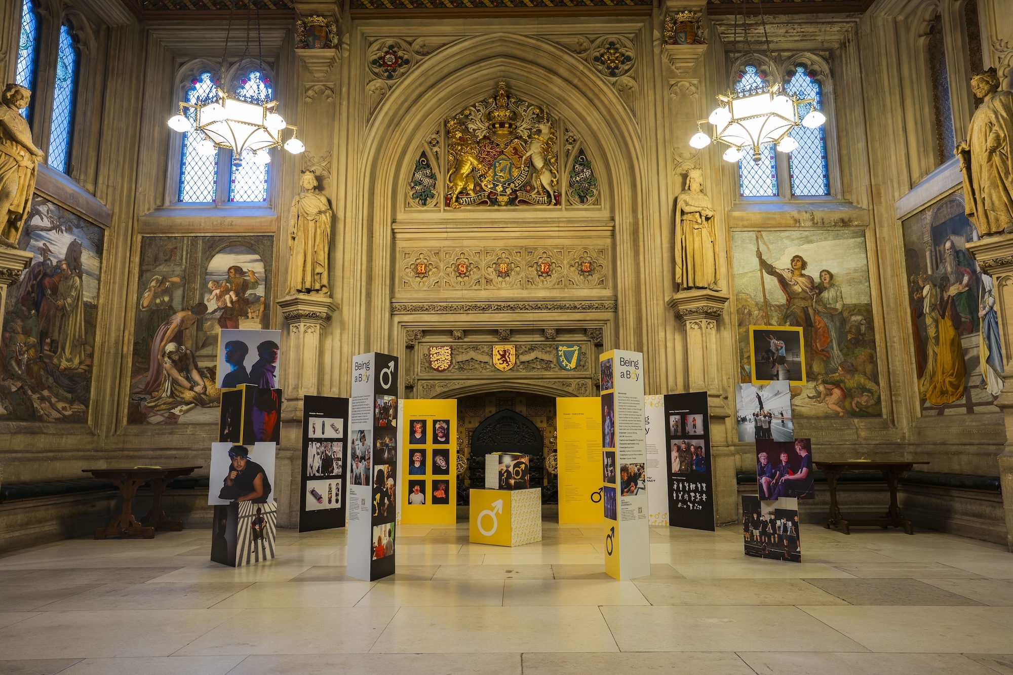Gothic hall with ornate golden arches, stained glass windows, and wall murals. Modern black exhibition displays arranged around central yellow booth on marble floor.