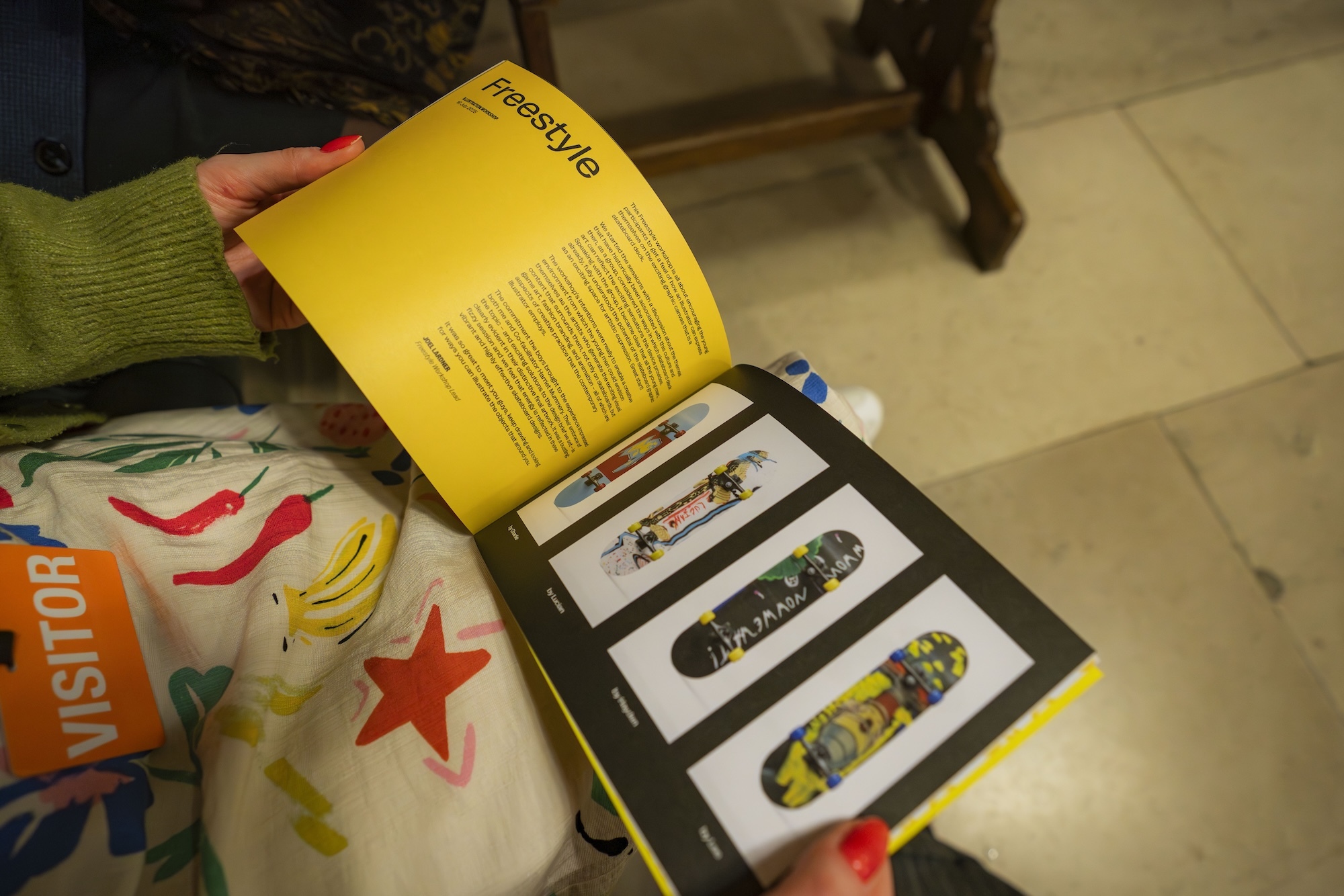 Person in green jumper holding open yellow and black book showing skateboard images, with colourful papers scattered on tiled floor.