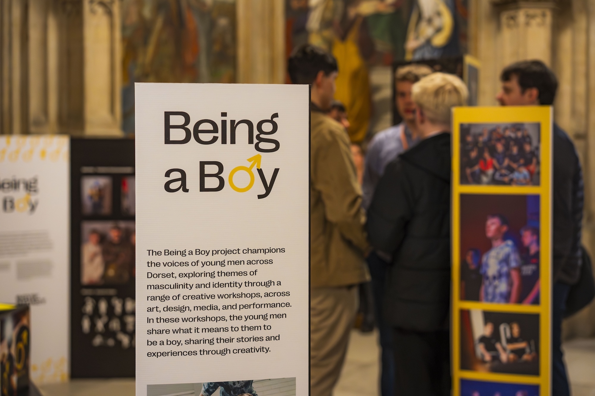 White exhibition banner with "Being a Boy" title in black text, yellow letter 'o'. People viewing colourful artwork displays in background.