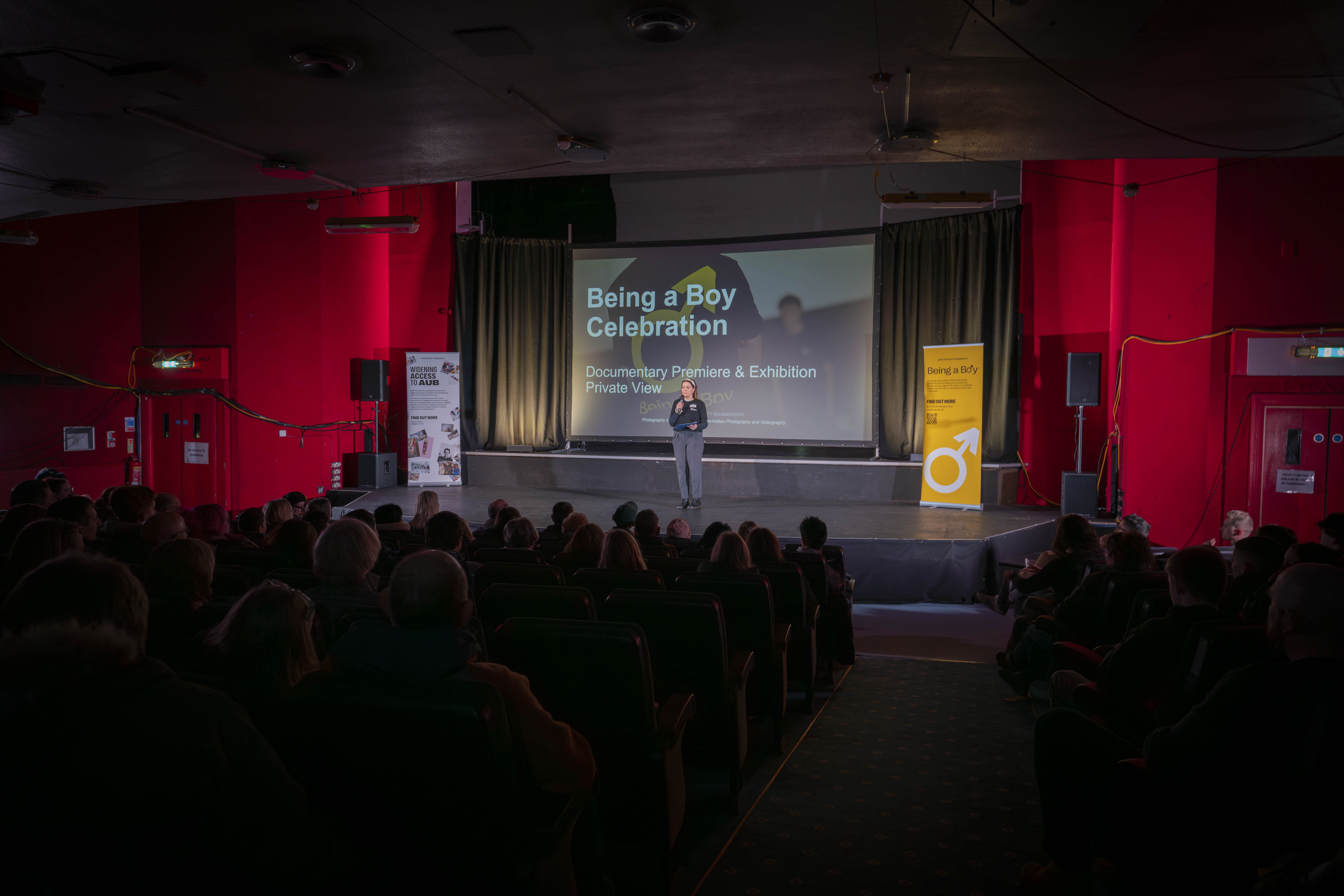 A member of staff on stage in front of a large screen welcoming guests to the Being a Boy Celebration