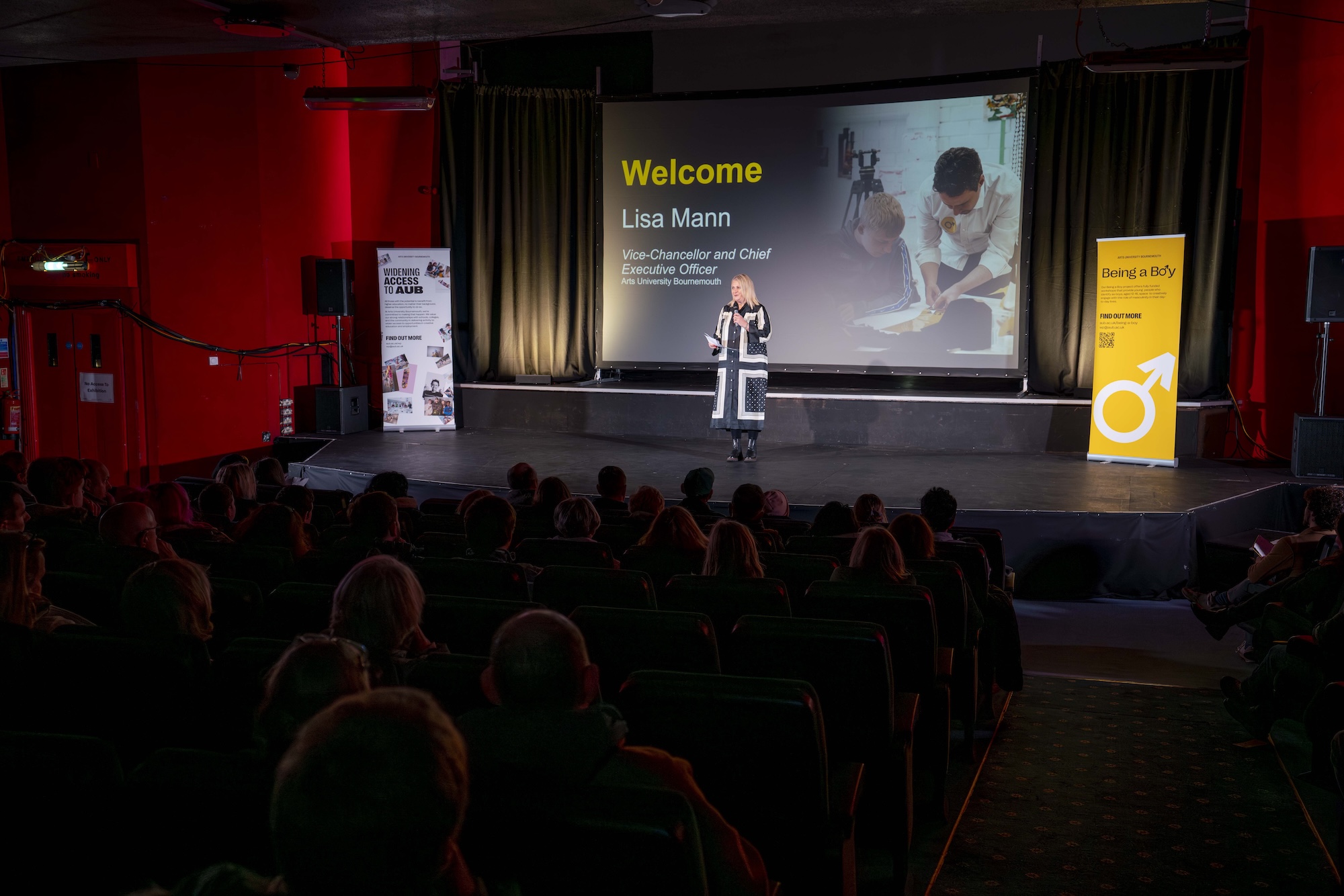 L Mann stands on a stage beside a screen with an audience seated in a dark theatre hall and banners on either side.