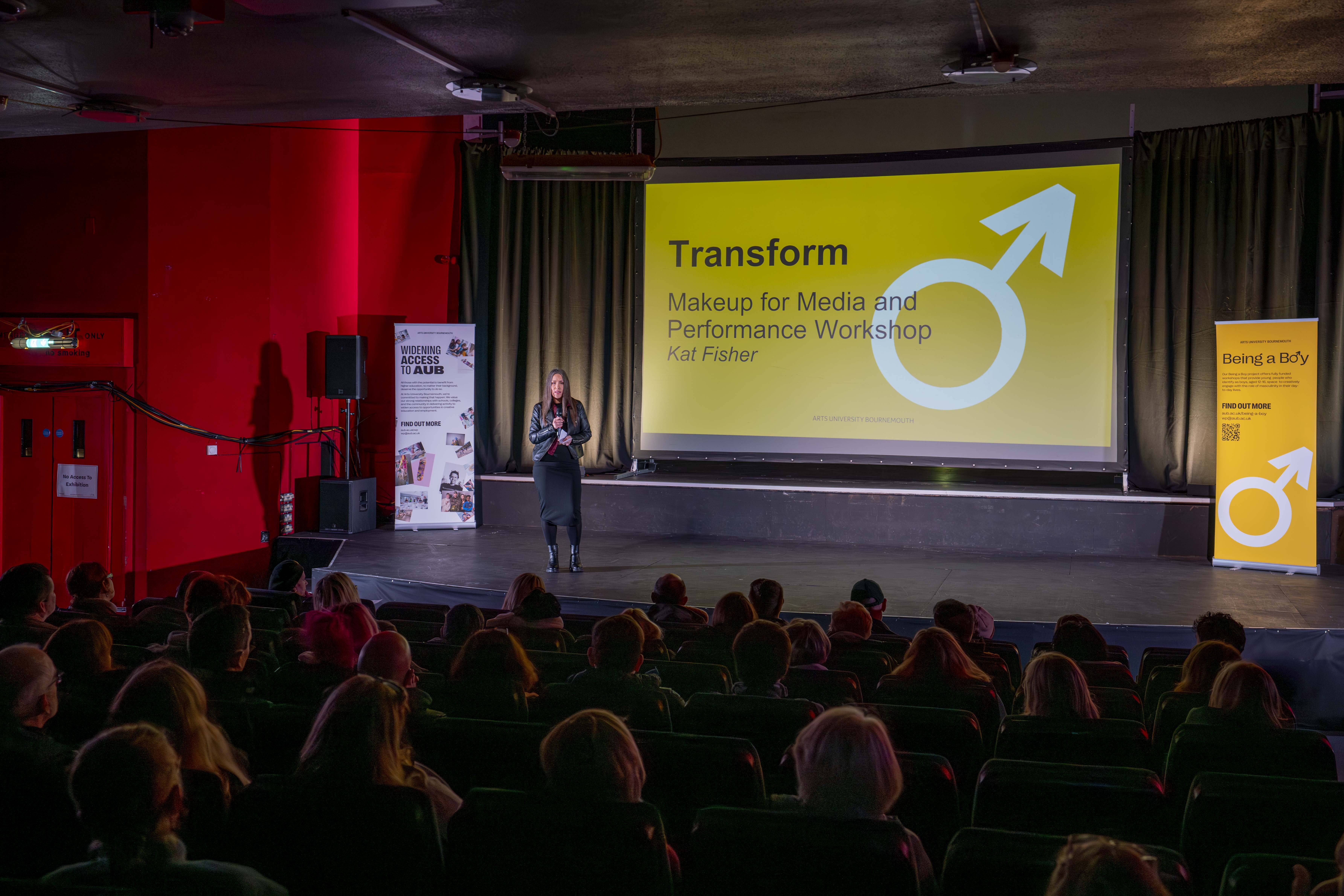Speaker presents to seated audience in conference hall with yellow "Transform" slide featuring blue arrow and male symbol on large screen.