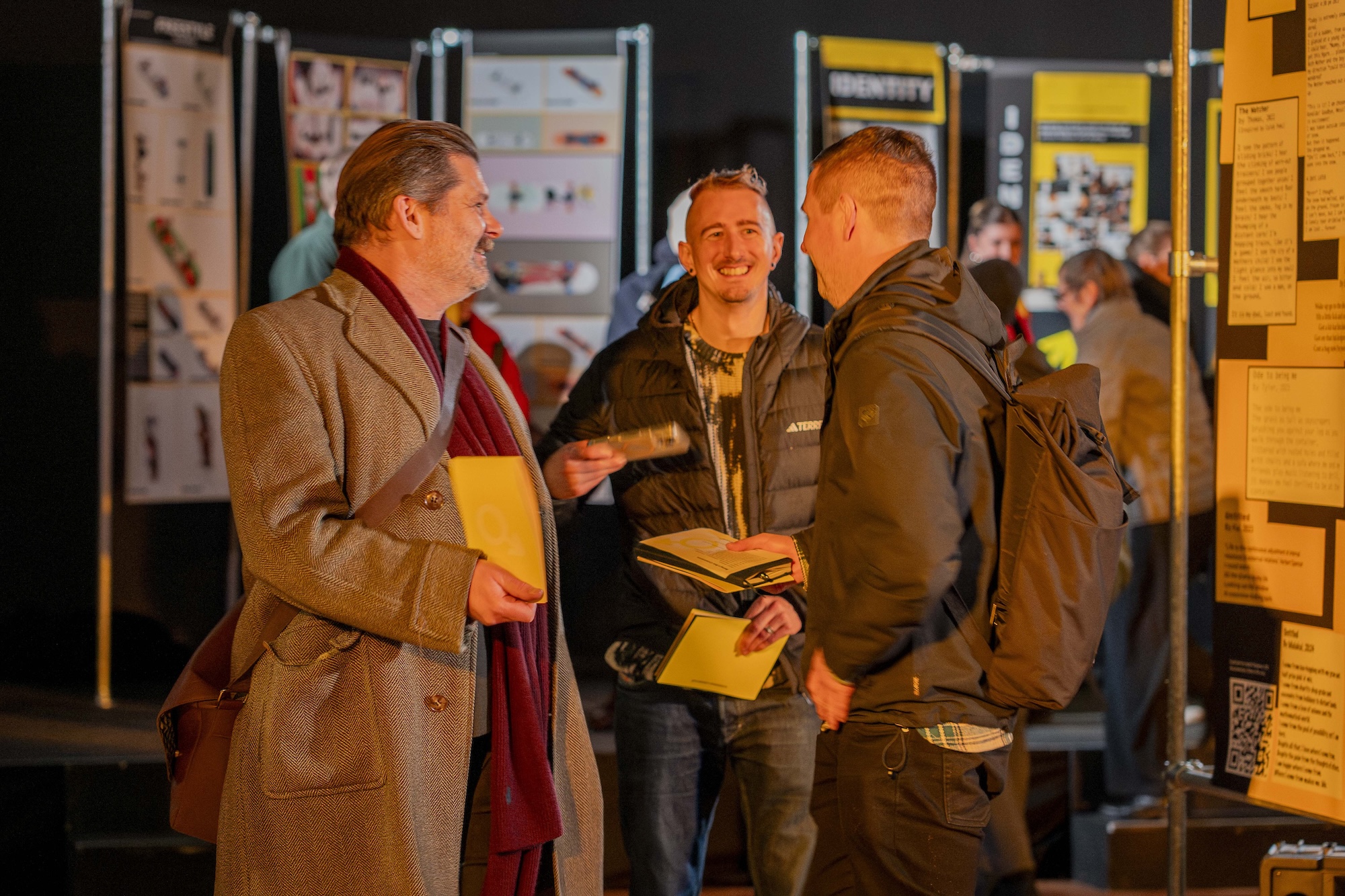 Three people talk in front of display boards at an indoor exhibition, with one holding papers and another wearing a backpack.