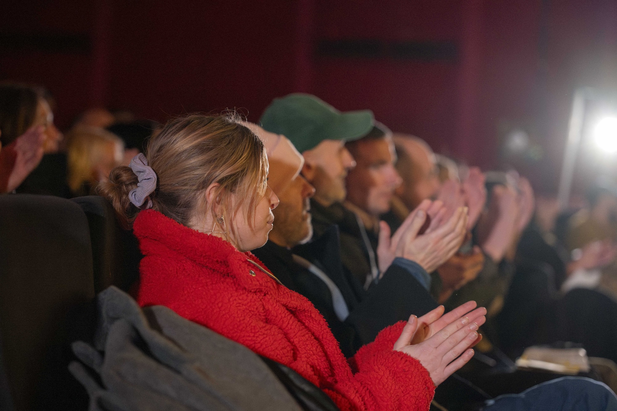 A person in a bright red coat sits among an audience, clapping in a dimly lit auditorium.