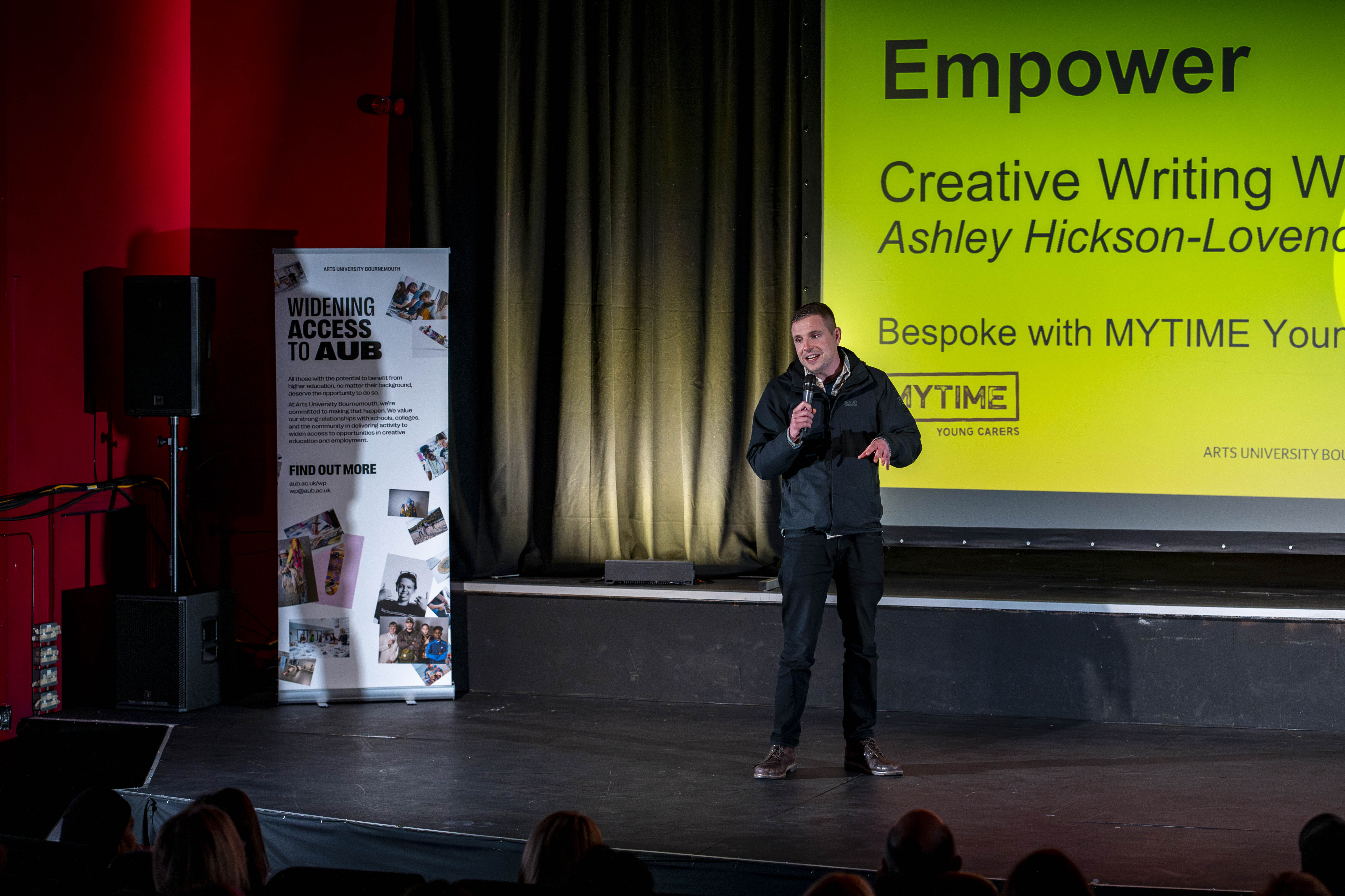 Alex Blower on a theatre stage, holding a microphone speaking