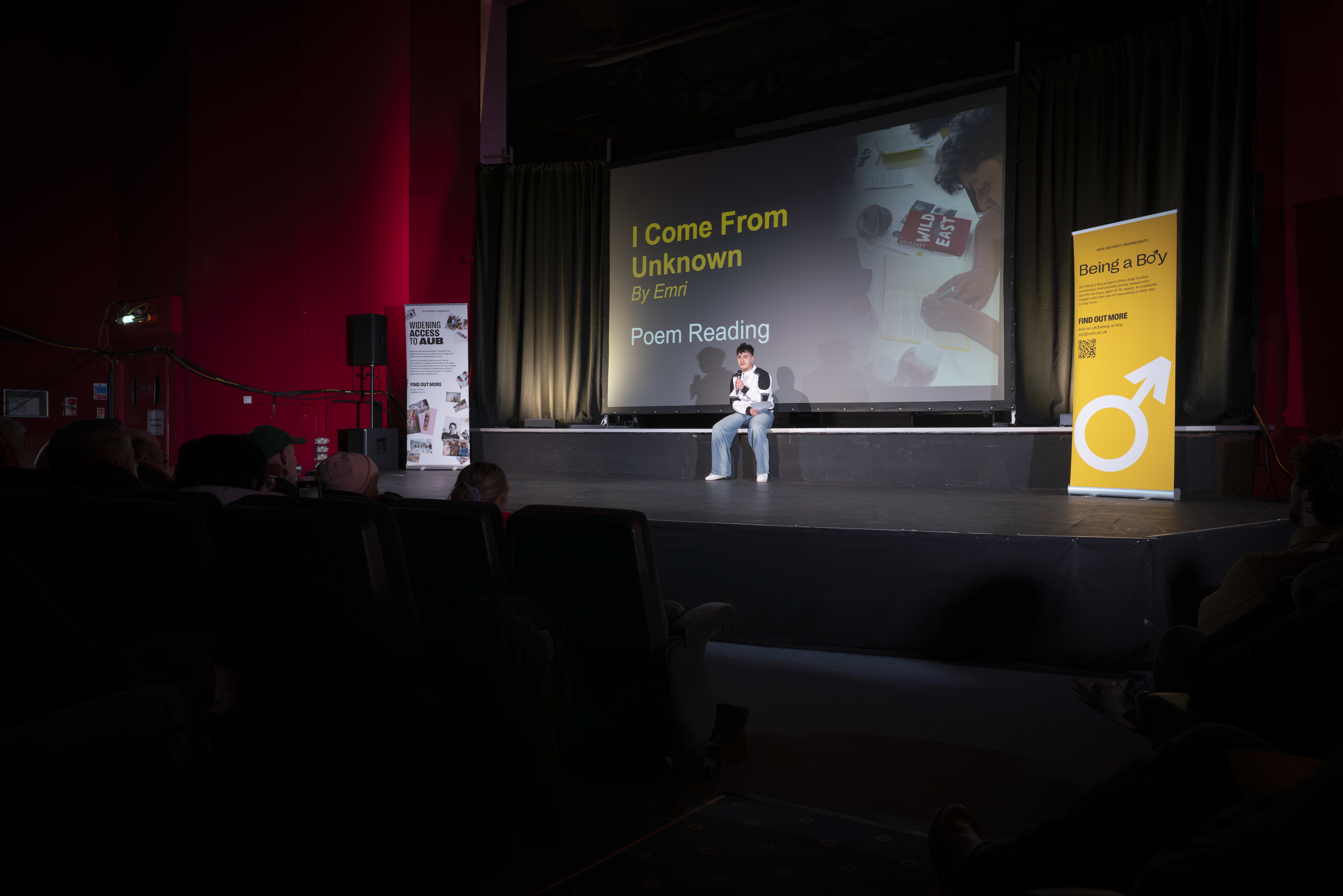 A young person sits alone on stage at the Palace Court Theatre, giving a poem reading. A screen behind them shows a slide with text reading "I Come From Unknown by Emri".