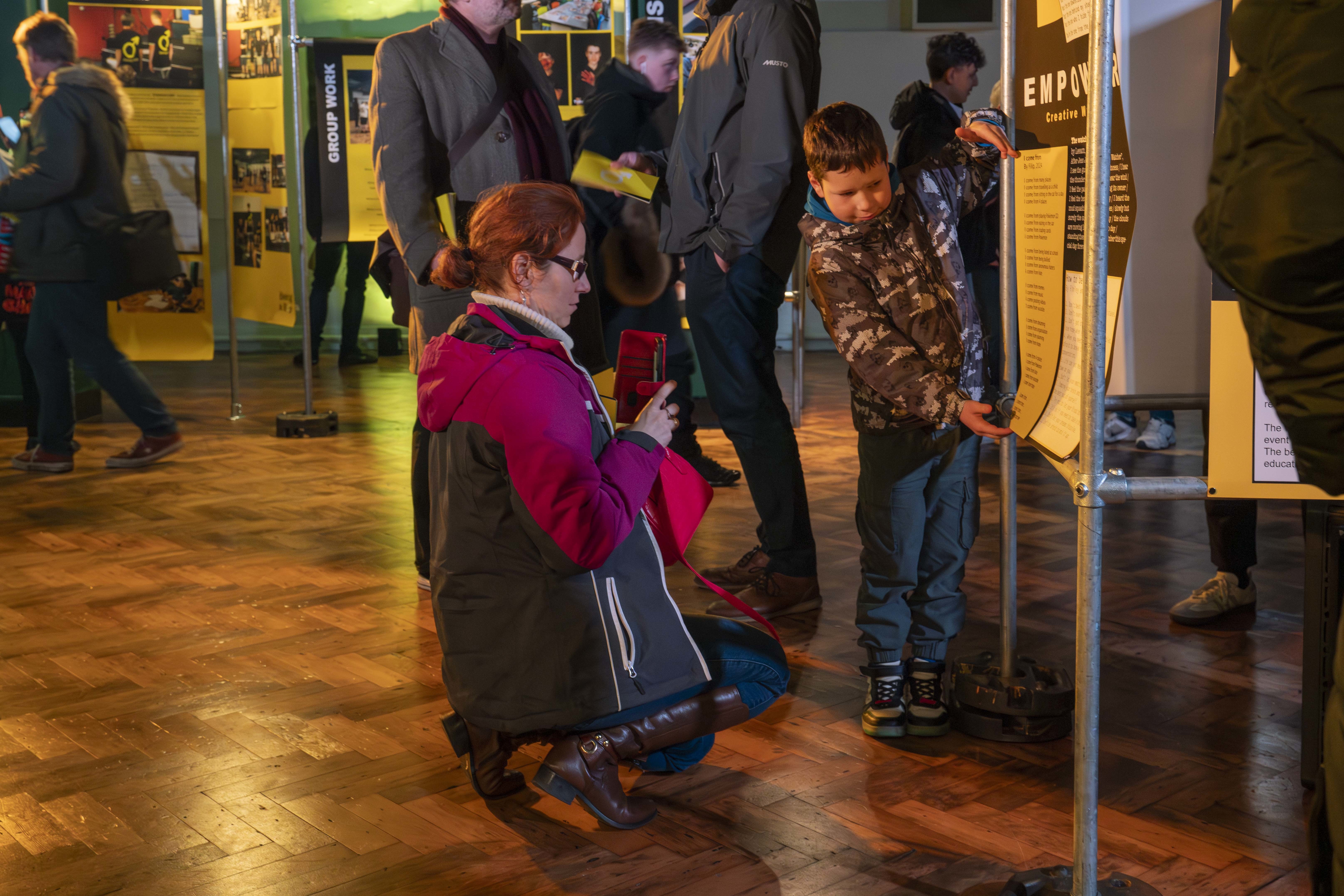 A young person presenting part of a poster in front of a parent who takes a photo on their phone.