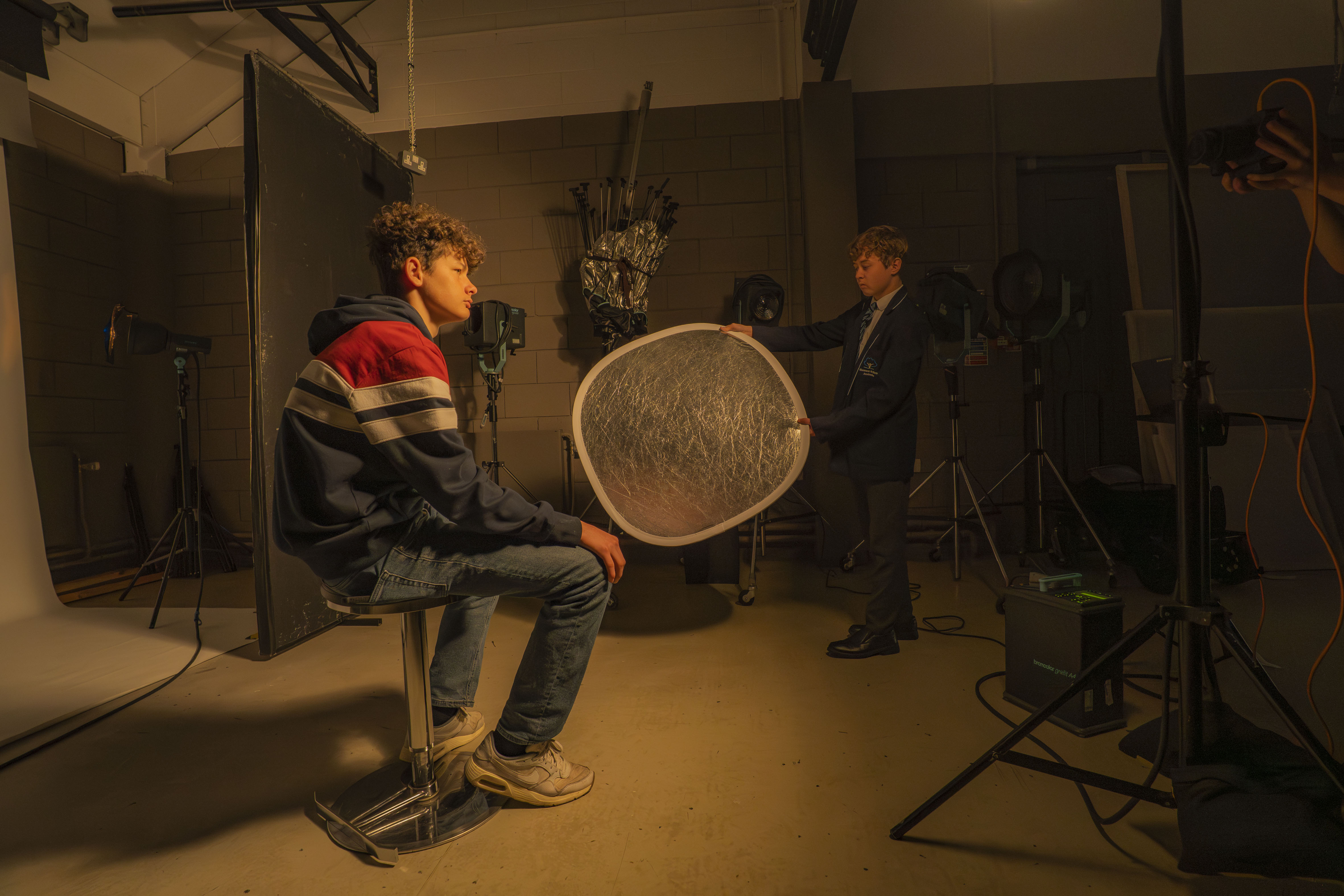 Two boys, one sat on a chair and the other holding a silver reflector under dim orange lighting in a photography studio.