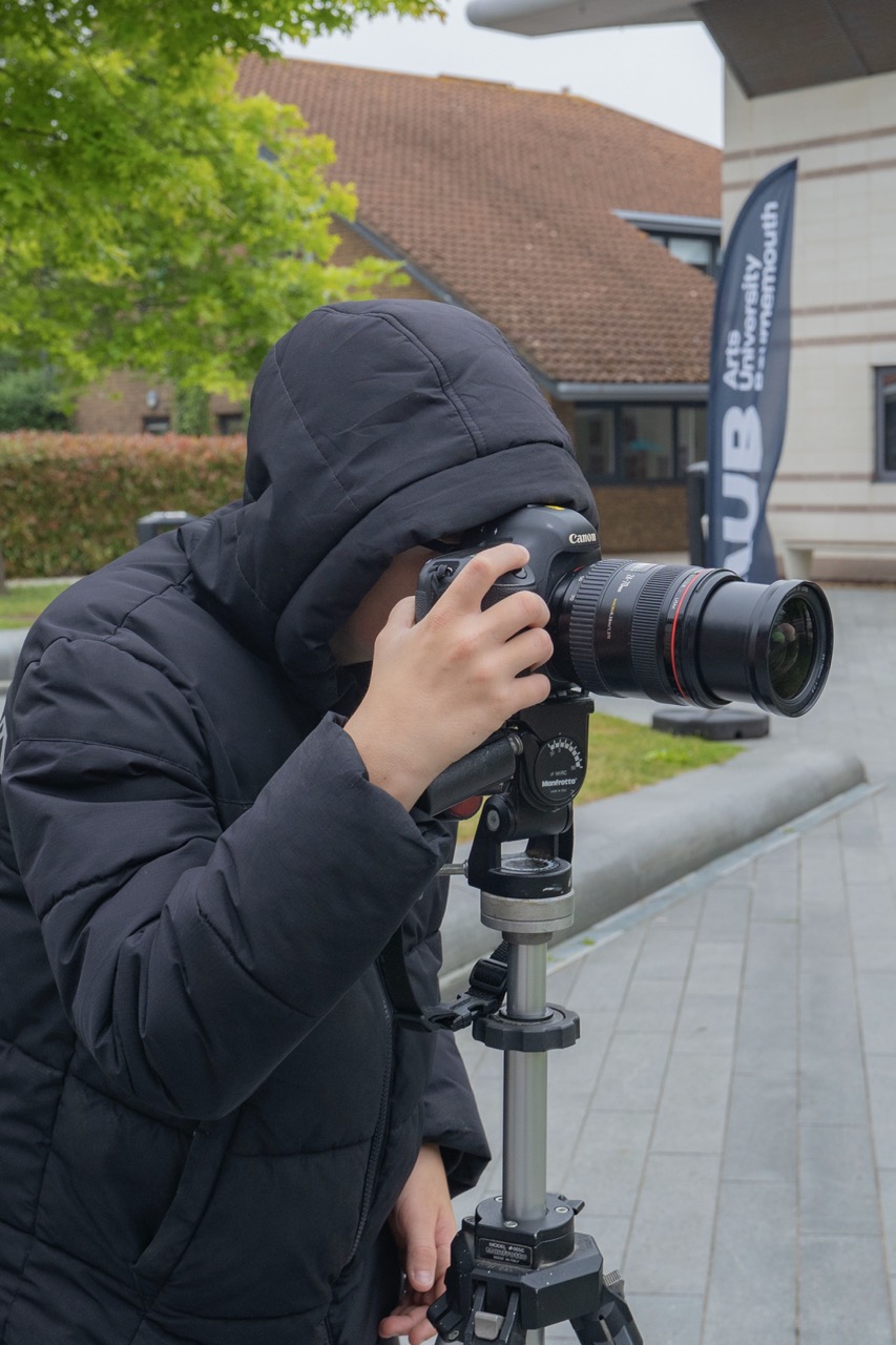 Person in thick black coat close to a camera on a tripod, face obscured, blurred trees and AUB campus in the background.