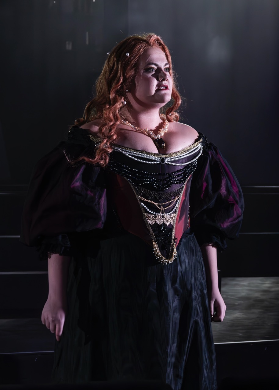 Woman in elaborate black period costume with ornate gold chain necklaces and purple cape, standing on stage steps with dramatic lighting.