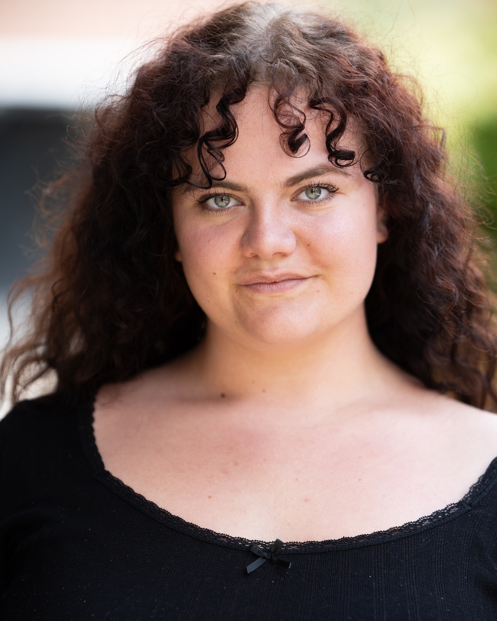 Woman with brown curly hair and blue eyes wearing black top, smiling at camera with soft outdoor lighting.