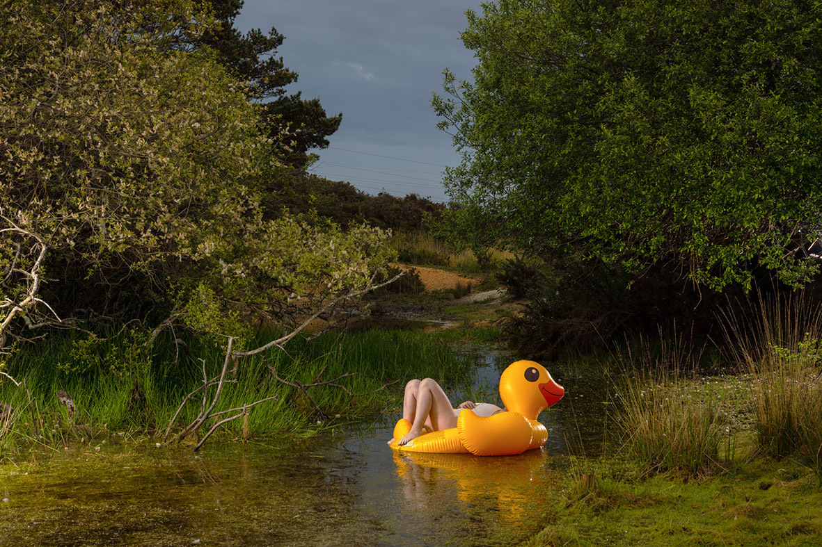 Person sitting on large yellow rubber duck in shallow water surrounded by green vegetation and trees under grey sky.