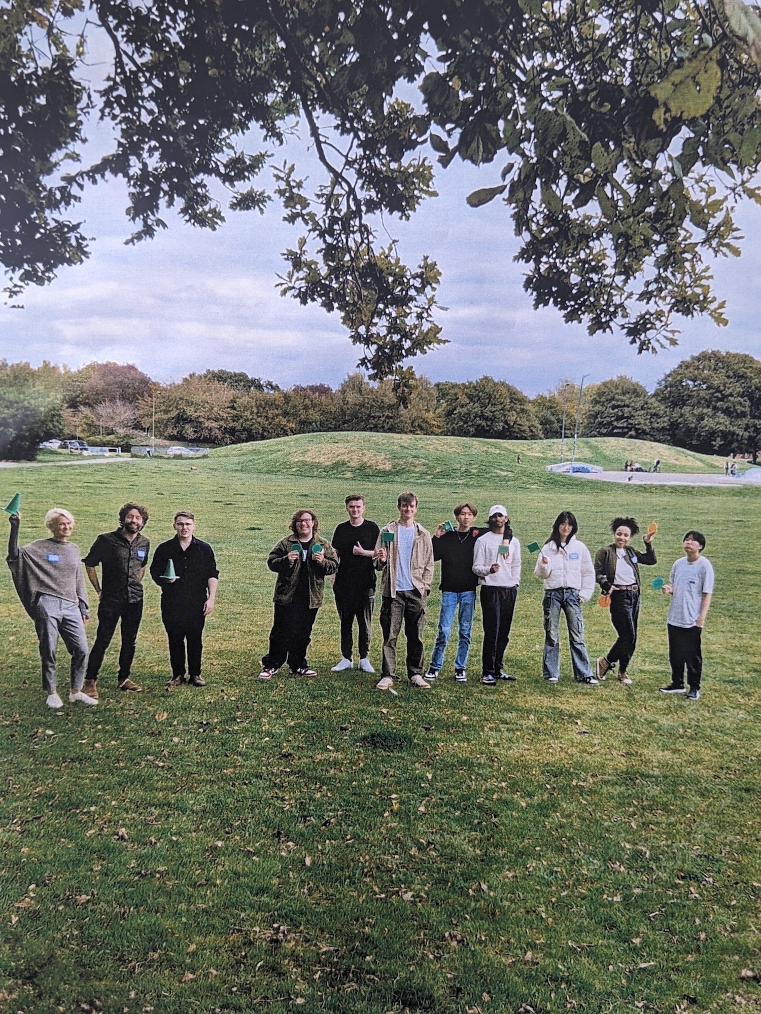 Group of 11 BA (Hons) Games Art and Design students and staff standing in a line on grass field under tree branches, with hills and cloudy sky in background.