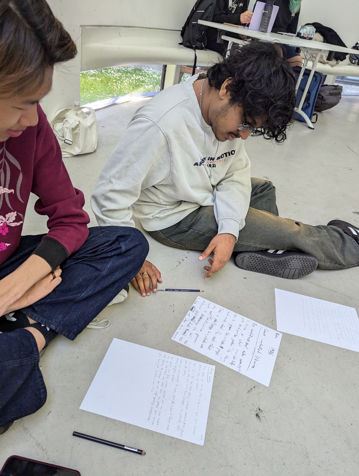 Two BA (Hons) Games Art and Design students sitting on classroom floor working with papers and pens spread between them, desks and bags visible in background.