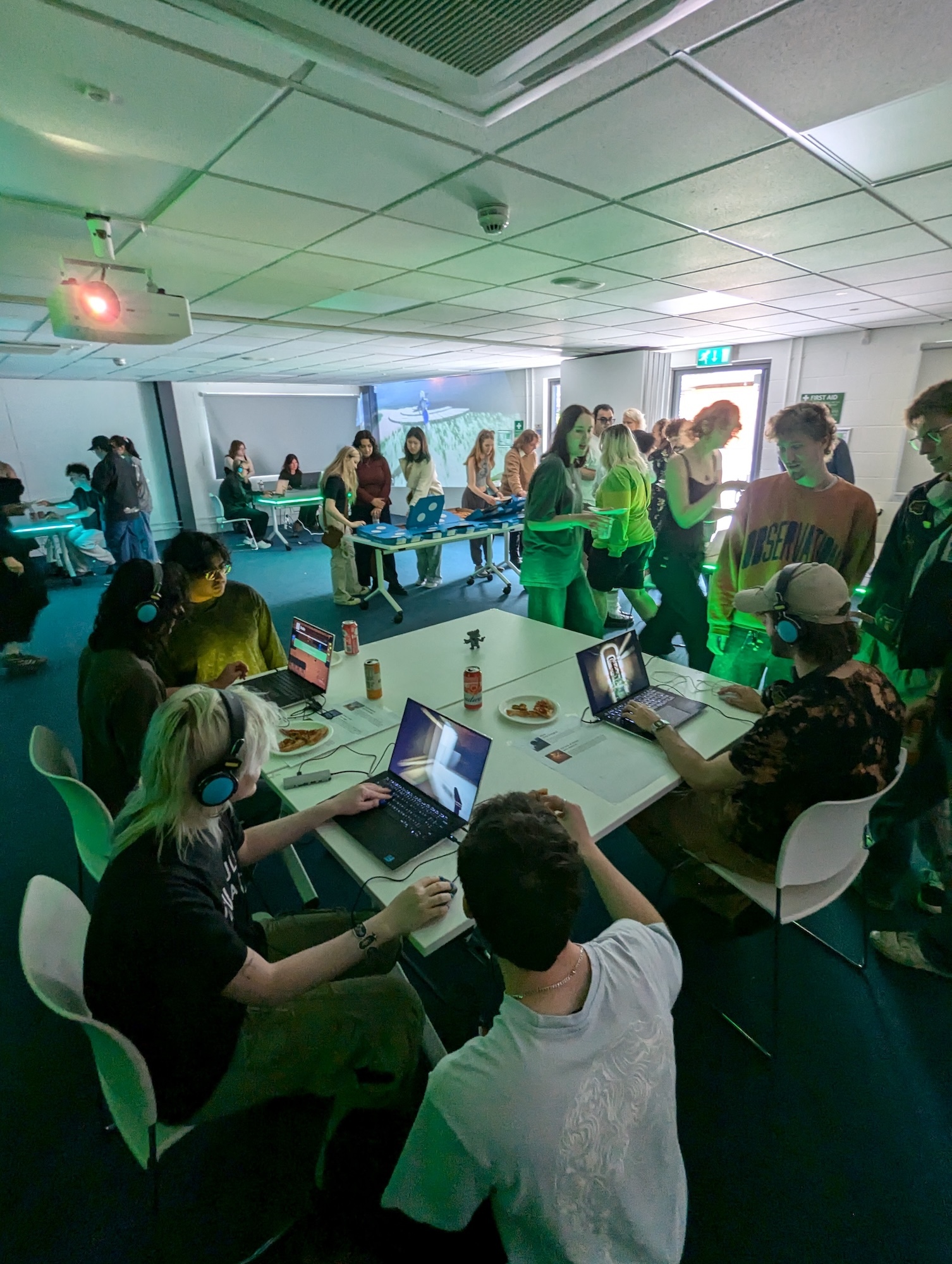 Conference Centre at Arts University Bournemouth with BA (Hons) Games Art and Design students and staff and visitors gathered around white tables using laptops at a games exhibition. Green and blue lighting illuminates white ceiling tiles and walls.
