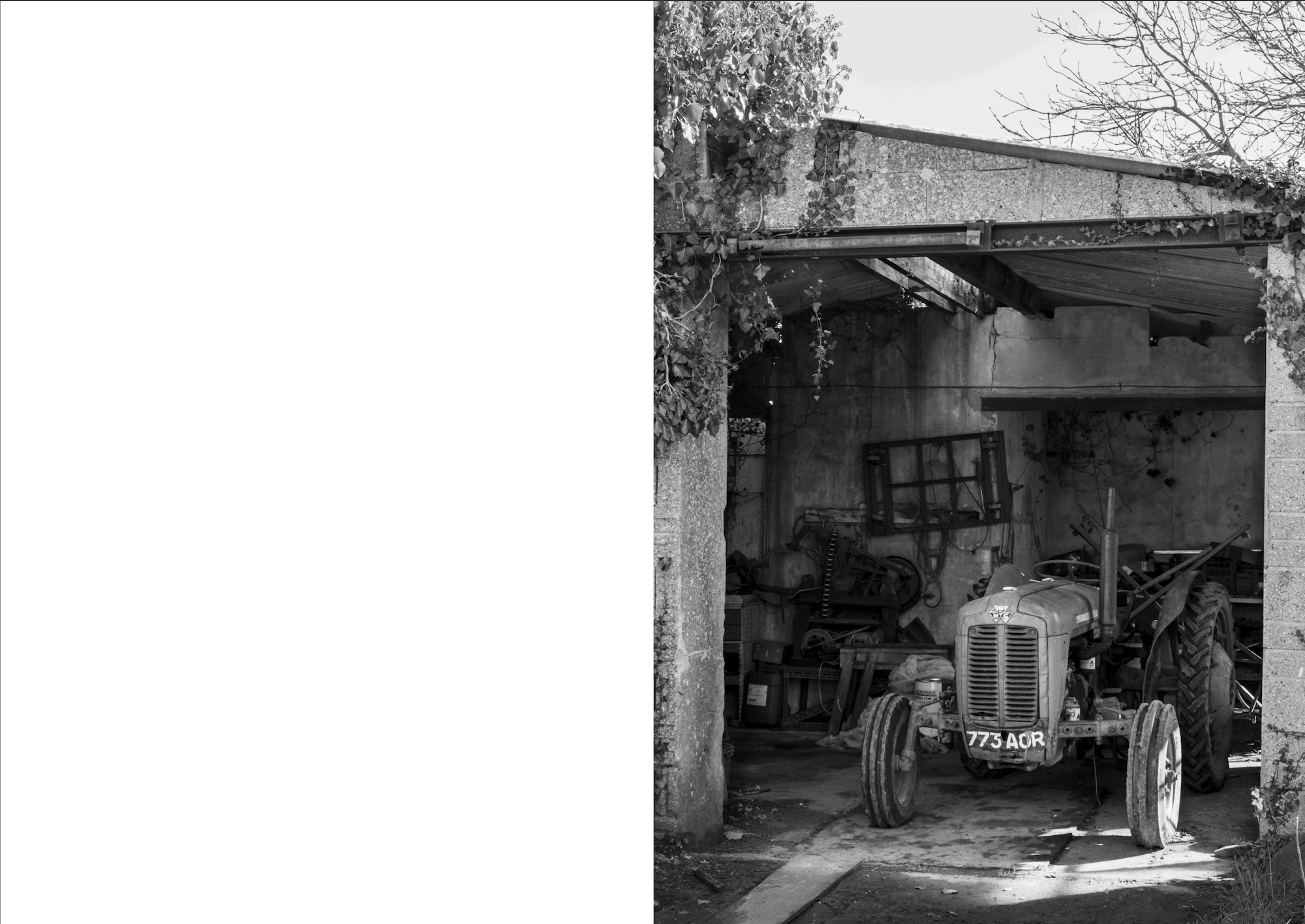 Black and white image showing vintage tractor parked in wooden garage or shed with corrugated roof and bare trees visible outside.