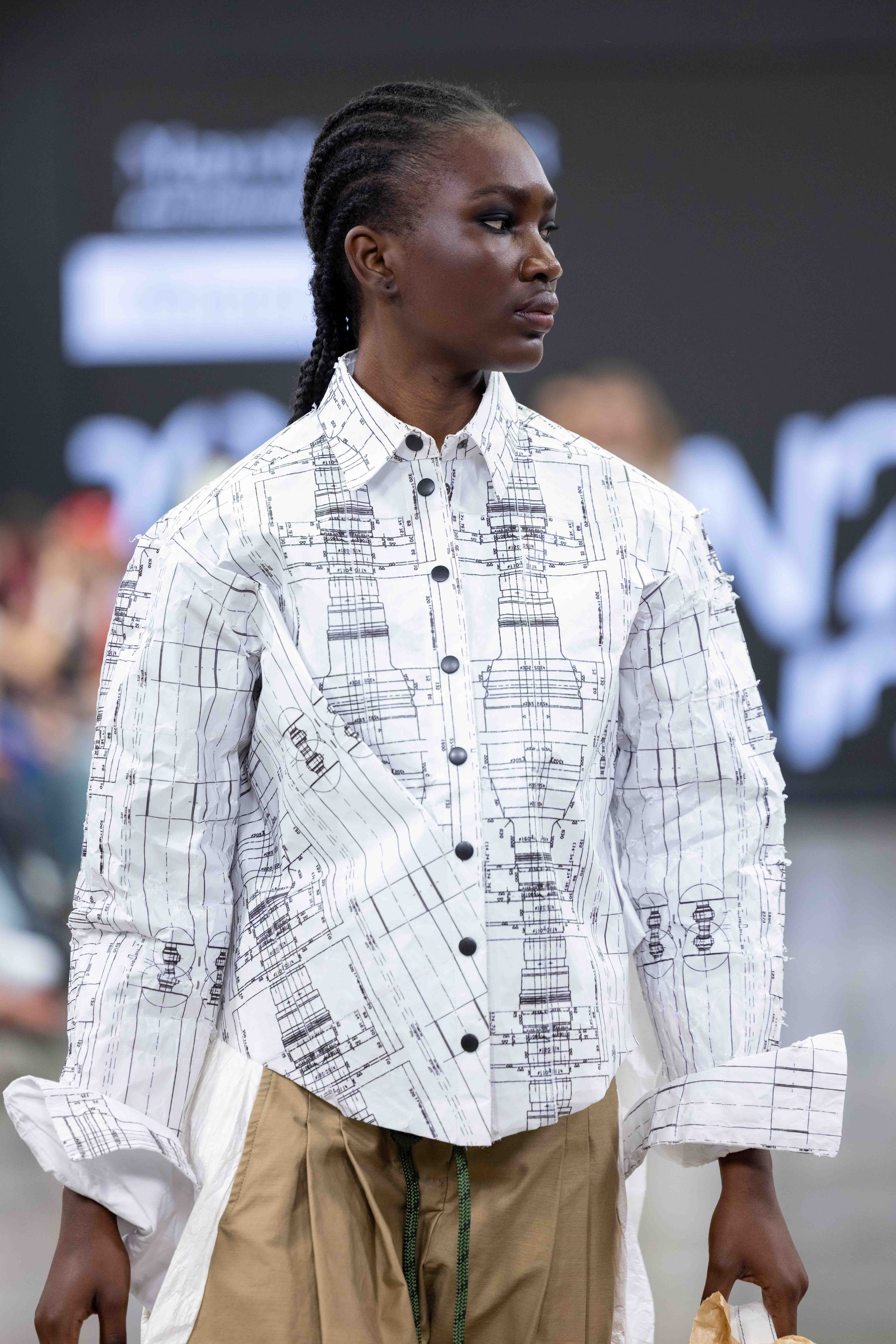 A model walks down the runway wearing a white button-up shirt printed with architectural drawings, paired with beige trousers. The background features a blurred audience and stage lighting.