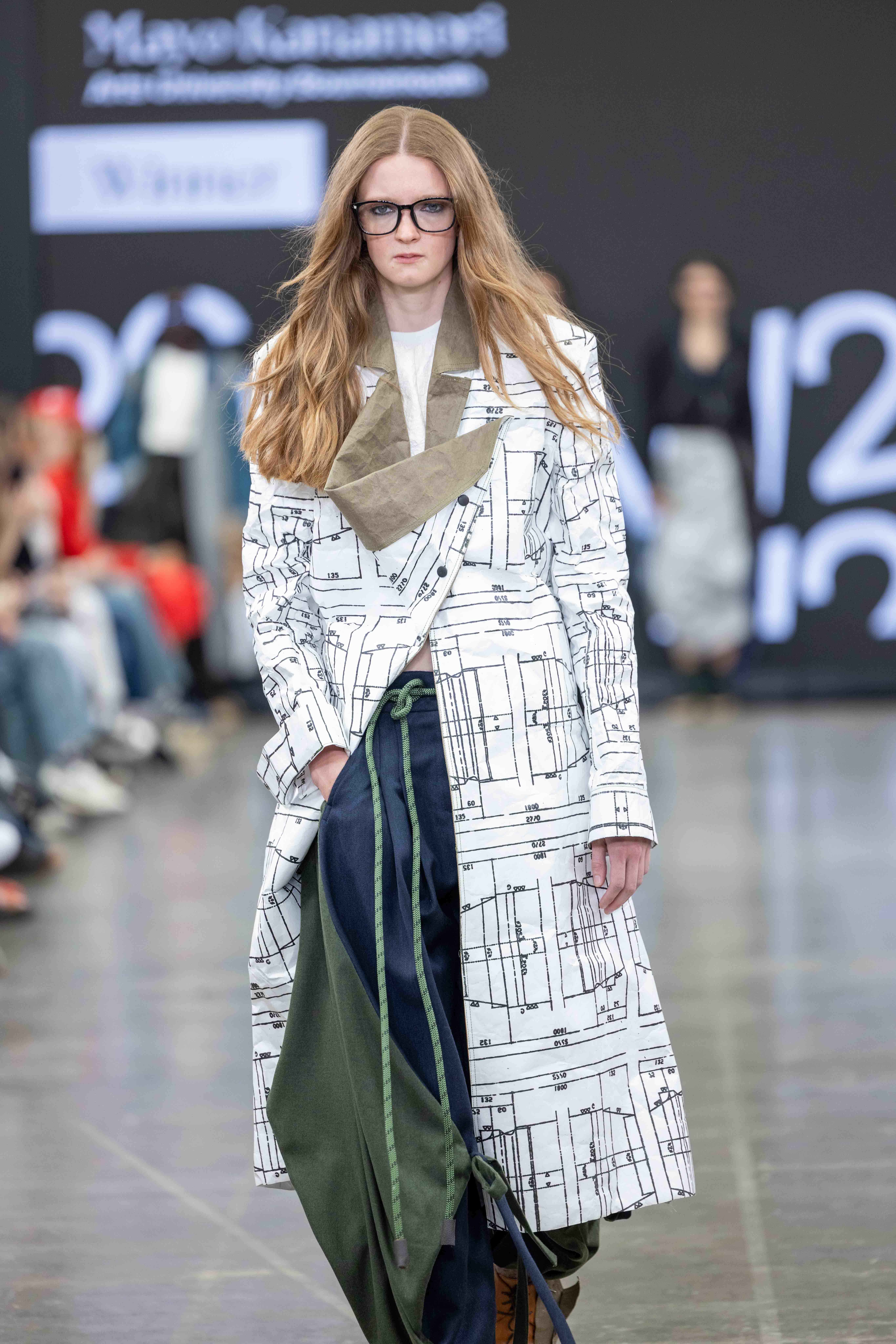 A model walks down the runway wearing a long white coat with a printed architectural design. She has long, wavy hair and glasses, and is holding a green garment, with a blue outfit visible below.