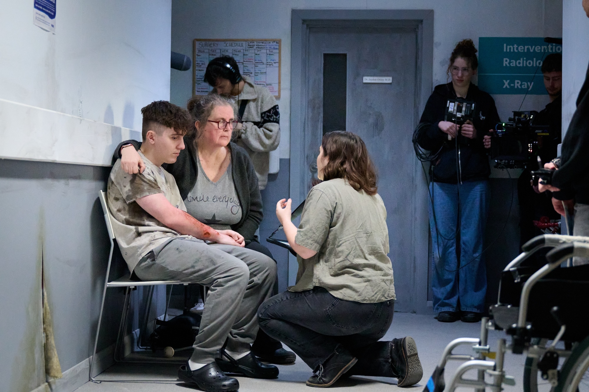 BA (Hons) Film Production student B Barr kneels down and directs two background artists, sitting on chairs in a hospital corridor.