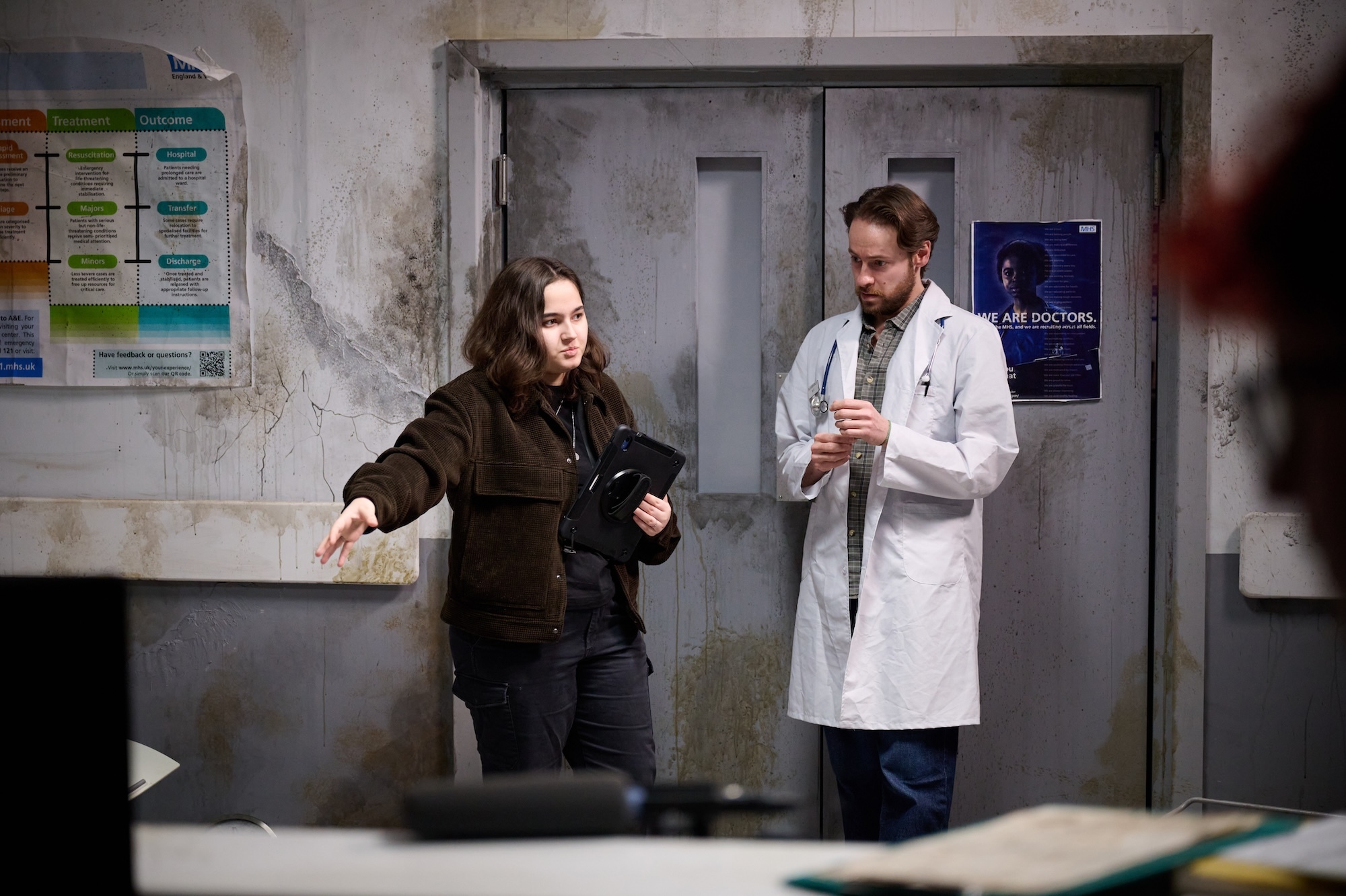 BA (Hons) Film Production student B Barr directing an actor in costume as a doctor, wearing a white lab coat, standing in front of a pair of hospital doors.