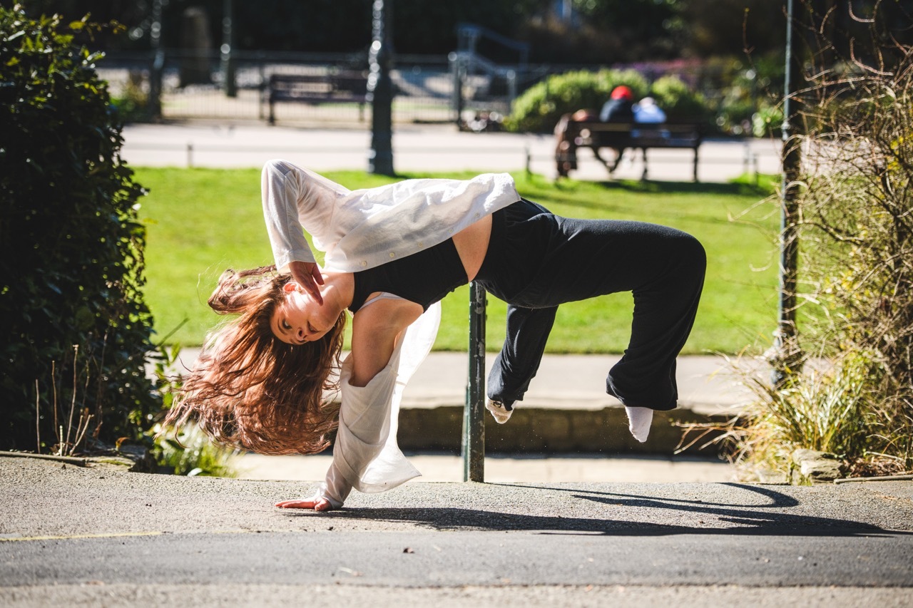 Woman performing freeze dance move on pavement, balancing horizontally on one hand with legs extended, park and trees in background.
