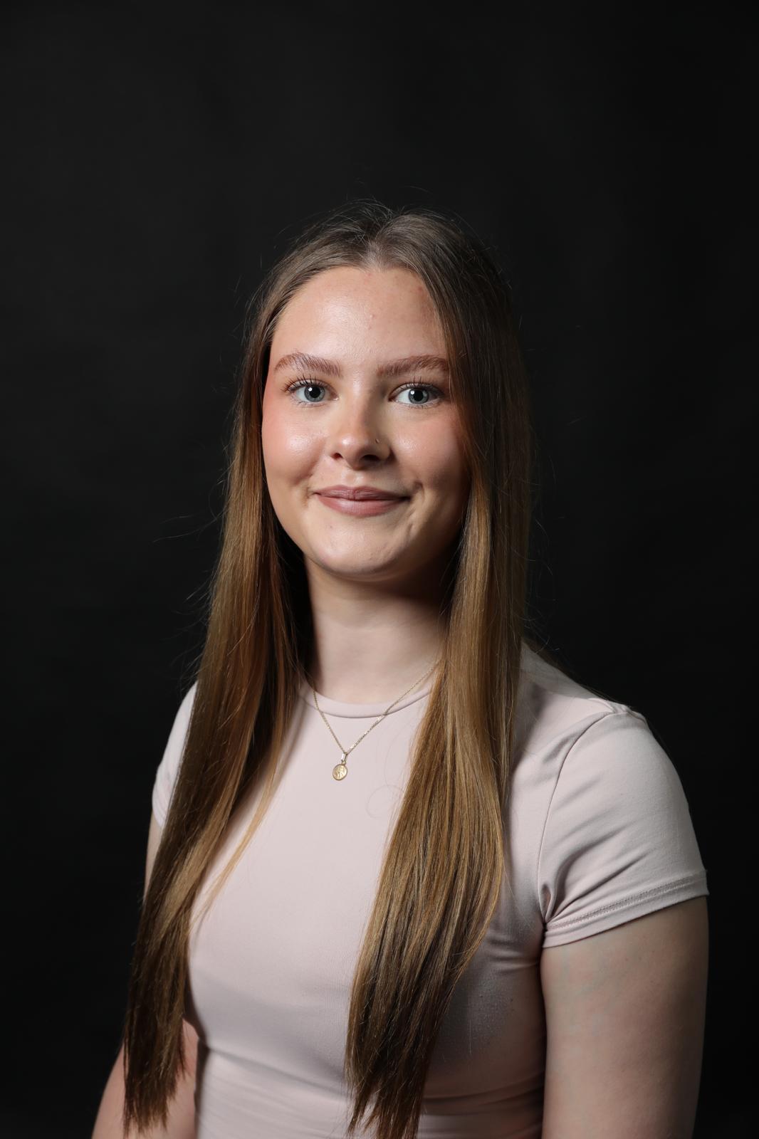 Young woman with long blonde hair wearing beige top and gold necklace, smiling against dark background.