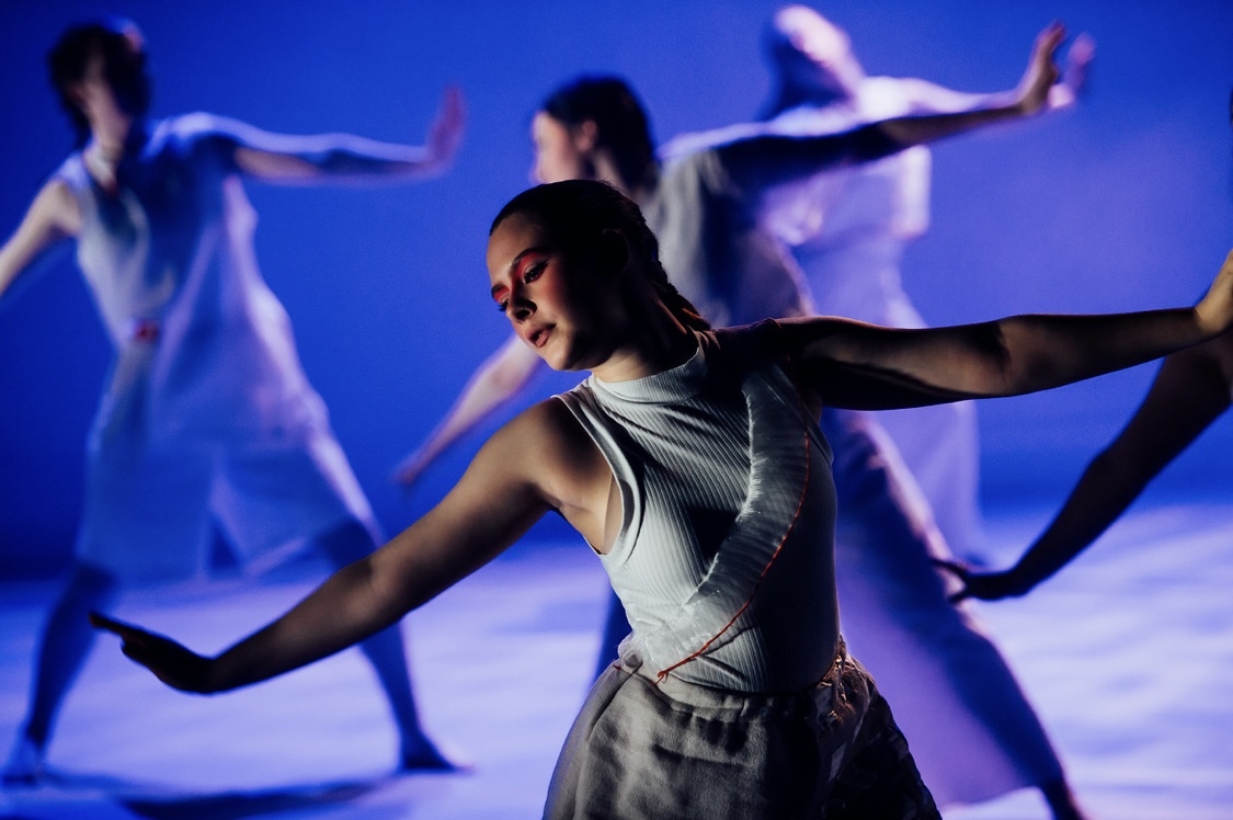 Dancer in grey top performs with arms extended whilst blurred figures move in background under blue and purple stage lighting.
