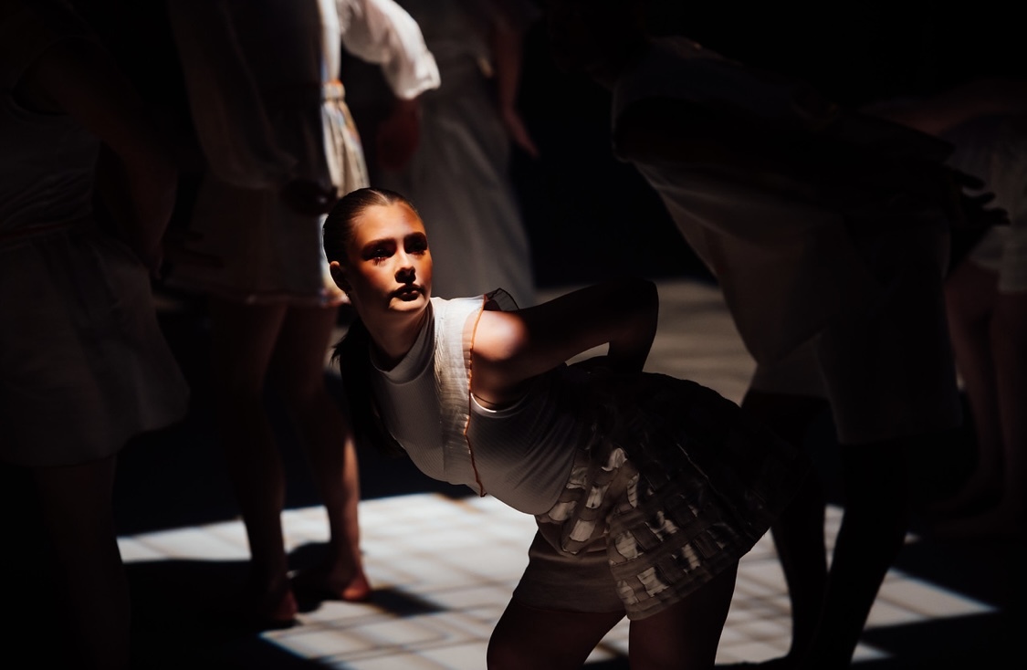 Dancer in white vest gesturing with raised arm, lit by dramatic spotlight against dark background with shadowy figures.