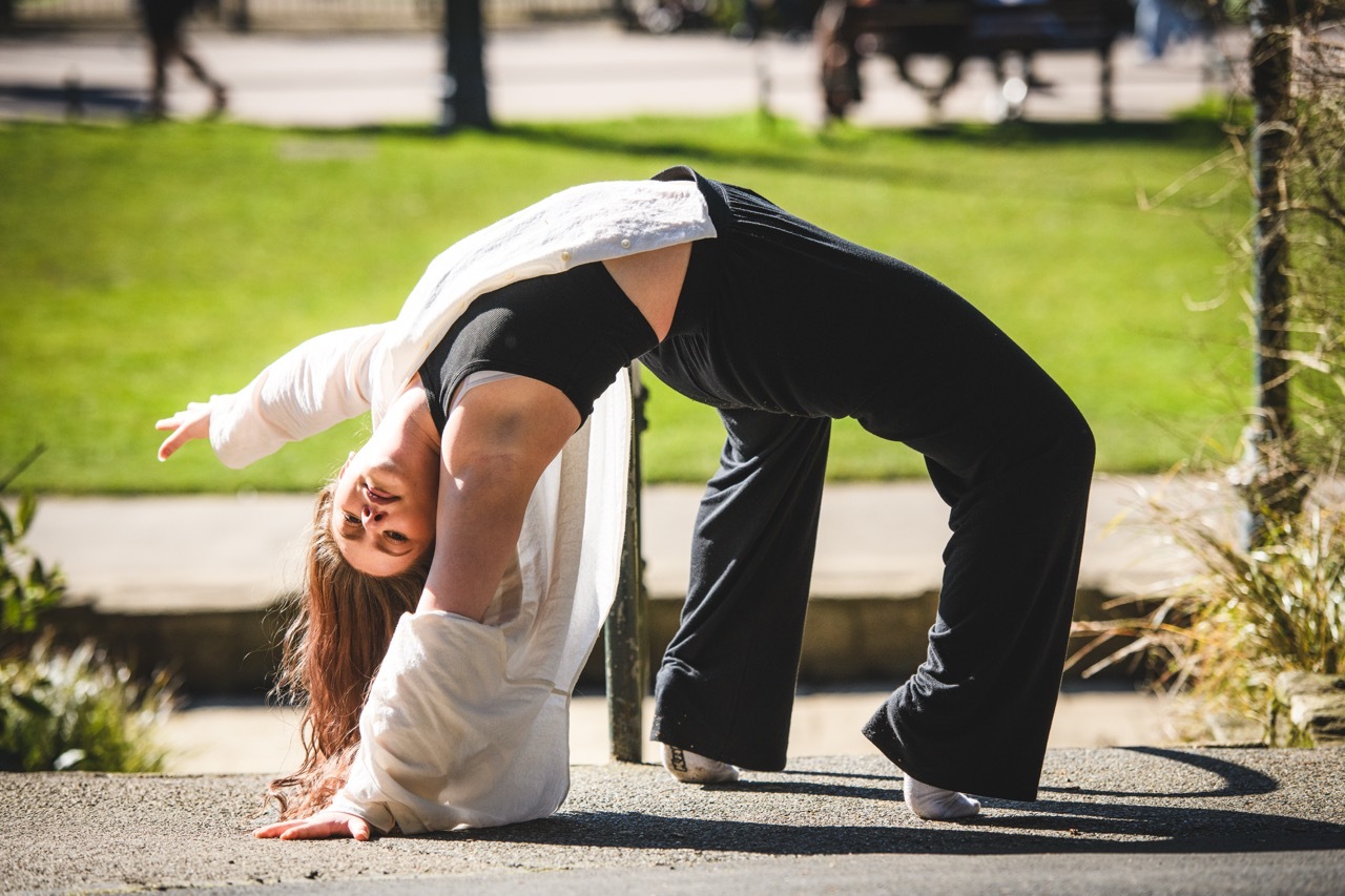 Woman in white top and black trousers performing backbend yoga pose on concrete surface with green grass and pathway in background.