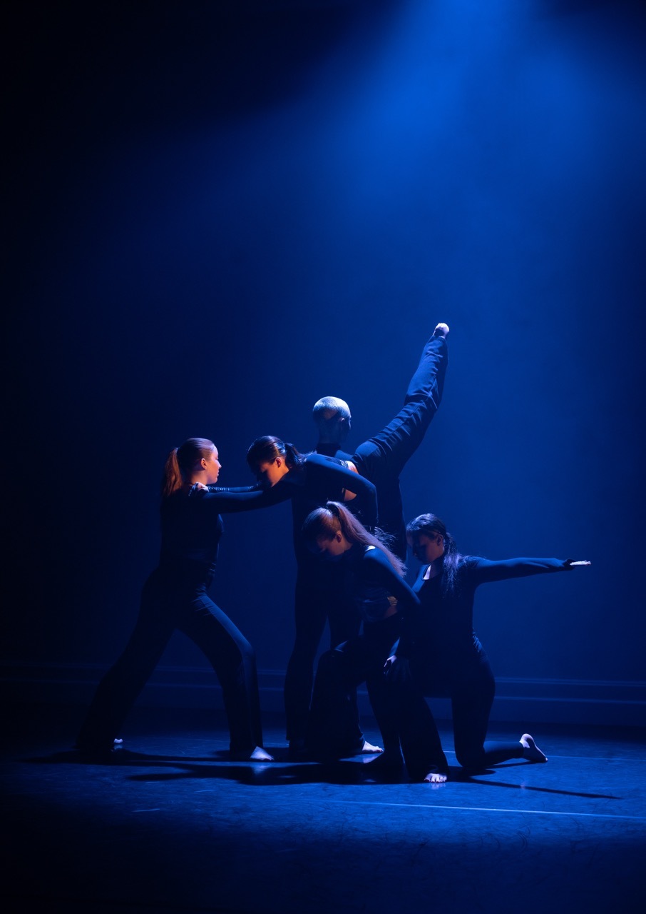 Five dancers in dark clothing performing on stage under blue lighting, with one dancer lifted horizontally above the others.