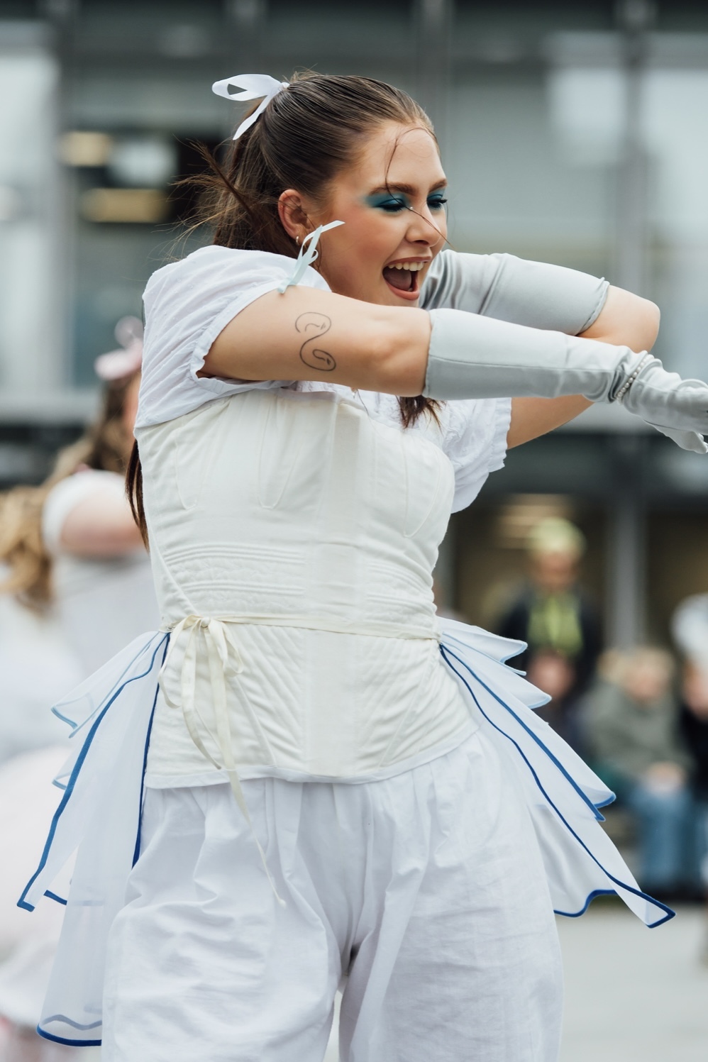 Woman in white dress with blue trim dancing outdoors, arms raised, mouth open, crowd watching in blurred background.