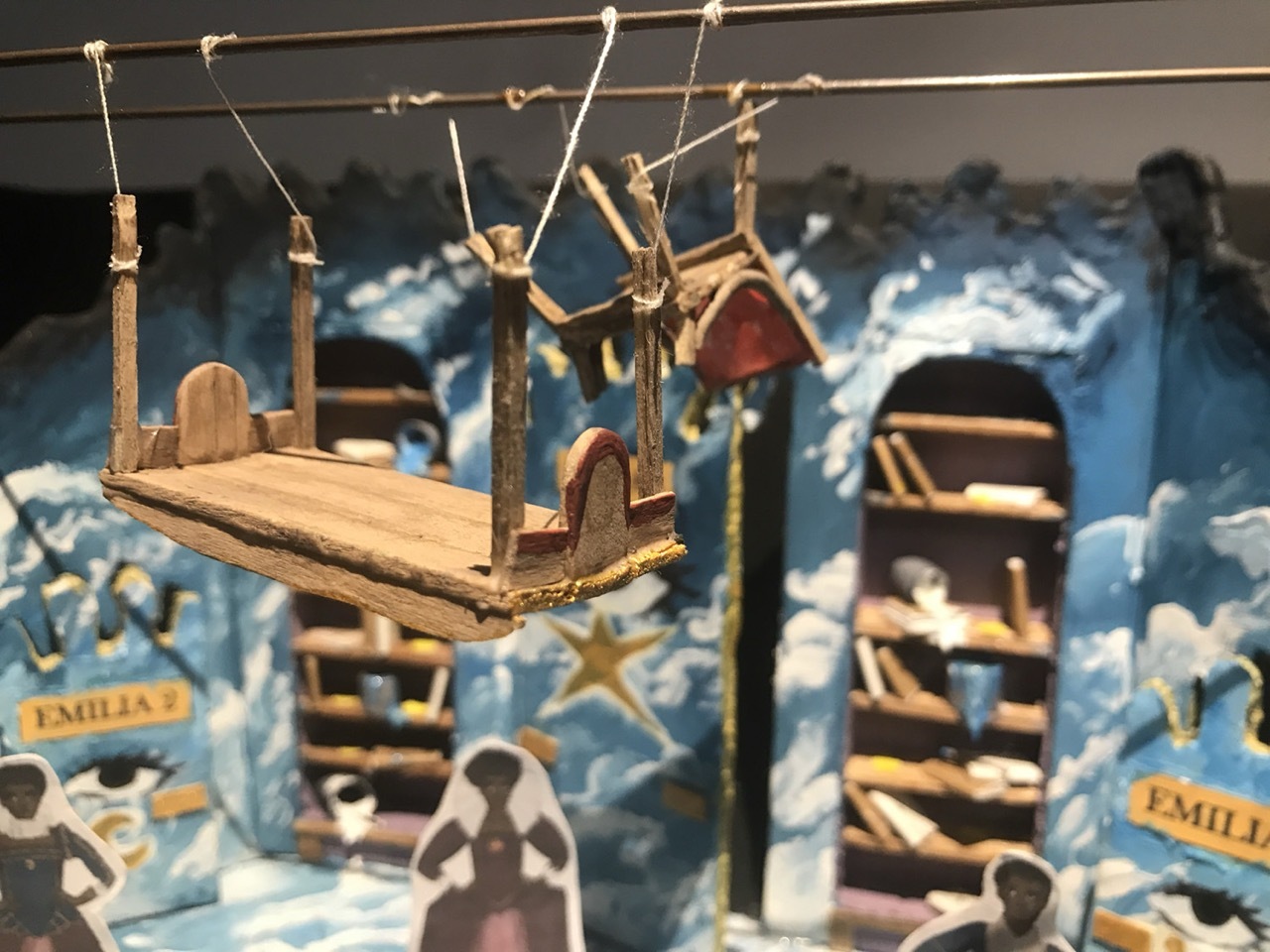 Miniature wooden sailing ship with masts and rigging displayed in glass case with blue maritime-themed background and other model boats.