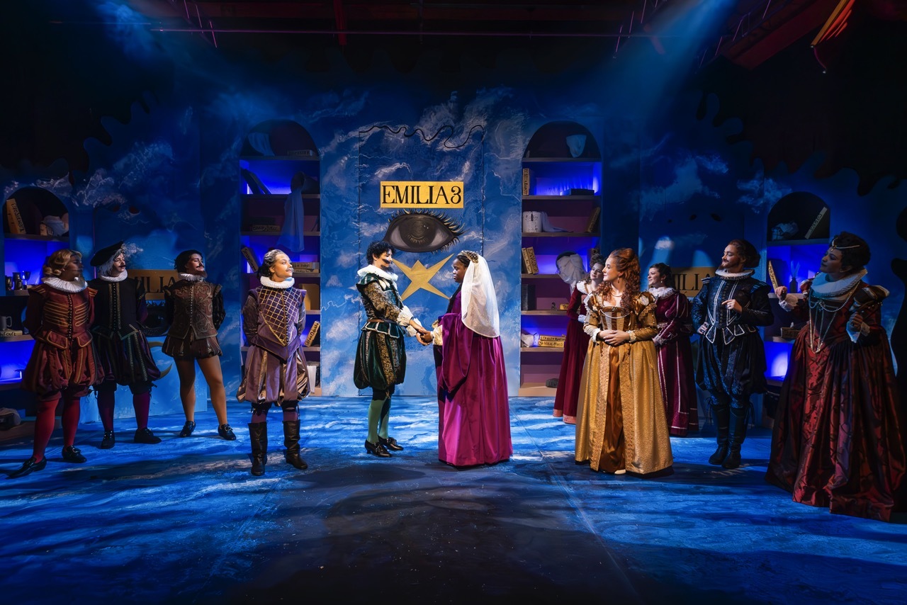 Theatre stage with performers in medieval costumes under blue lighting. Central figures in white and red robes, surrounded by armoured characters.