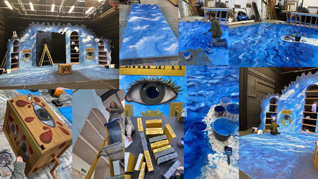 Large warehouse space with blue painted surfaces covering walls and floors, cardboard boxes, wooden structures, and geometric shapes scattered throughout.